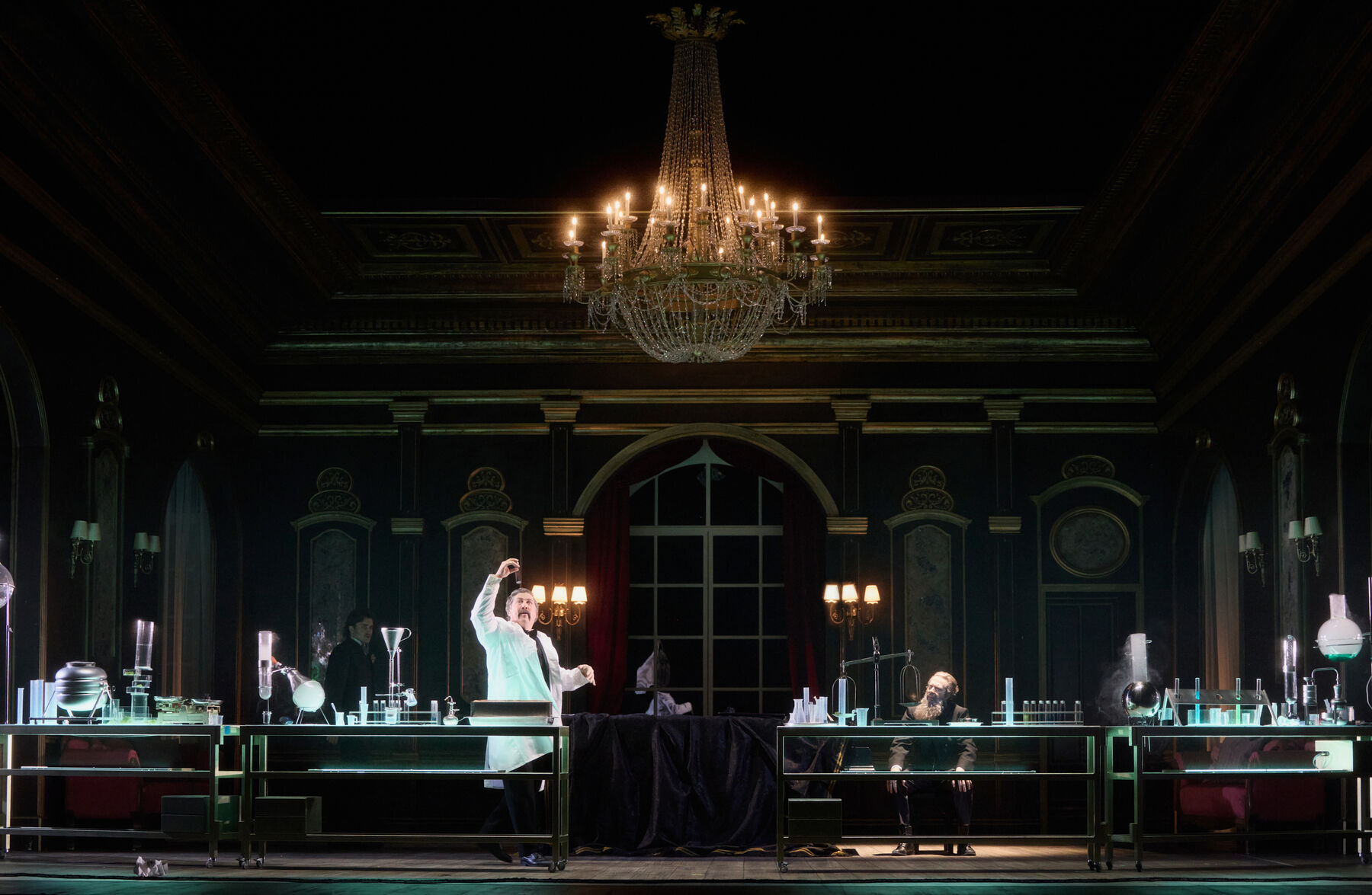 ein Chemielabor steht in einem prunkvollen Saal mit einem großen Kronleuchter und ein Flügel in der Mitte. Ein Mann im einem Laborkittel betrachtet ein Reagenzglas. Im Hintergrund sitzen zwei weitere Männer.