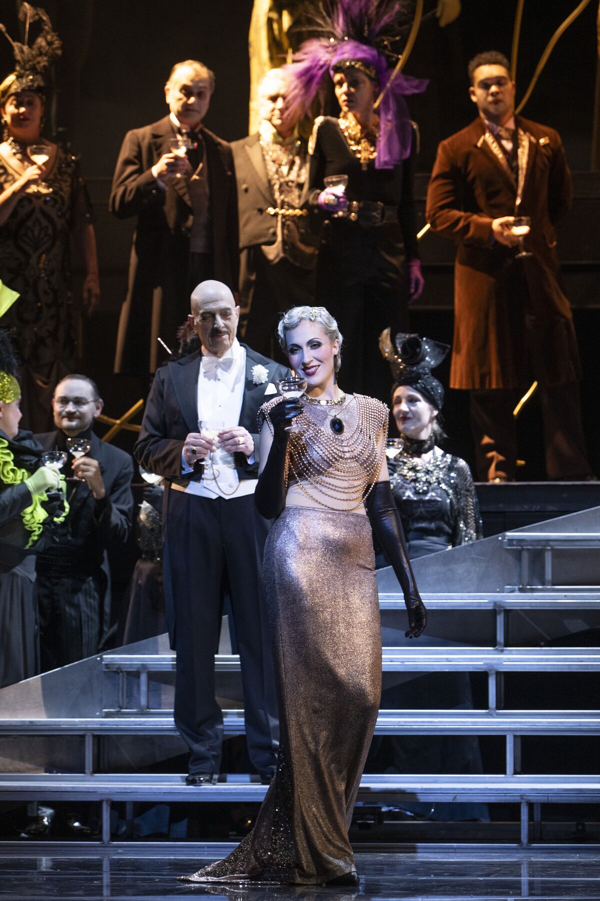 eine blonde Frau mit einem engen, silbernen Kleid steht mit einem Glas in der Hand, vor mehreren festlich gekleideten Menschen.