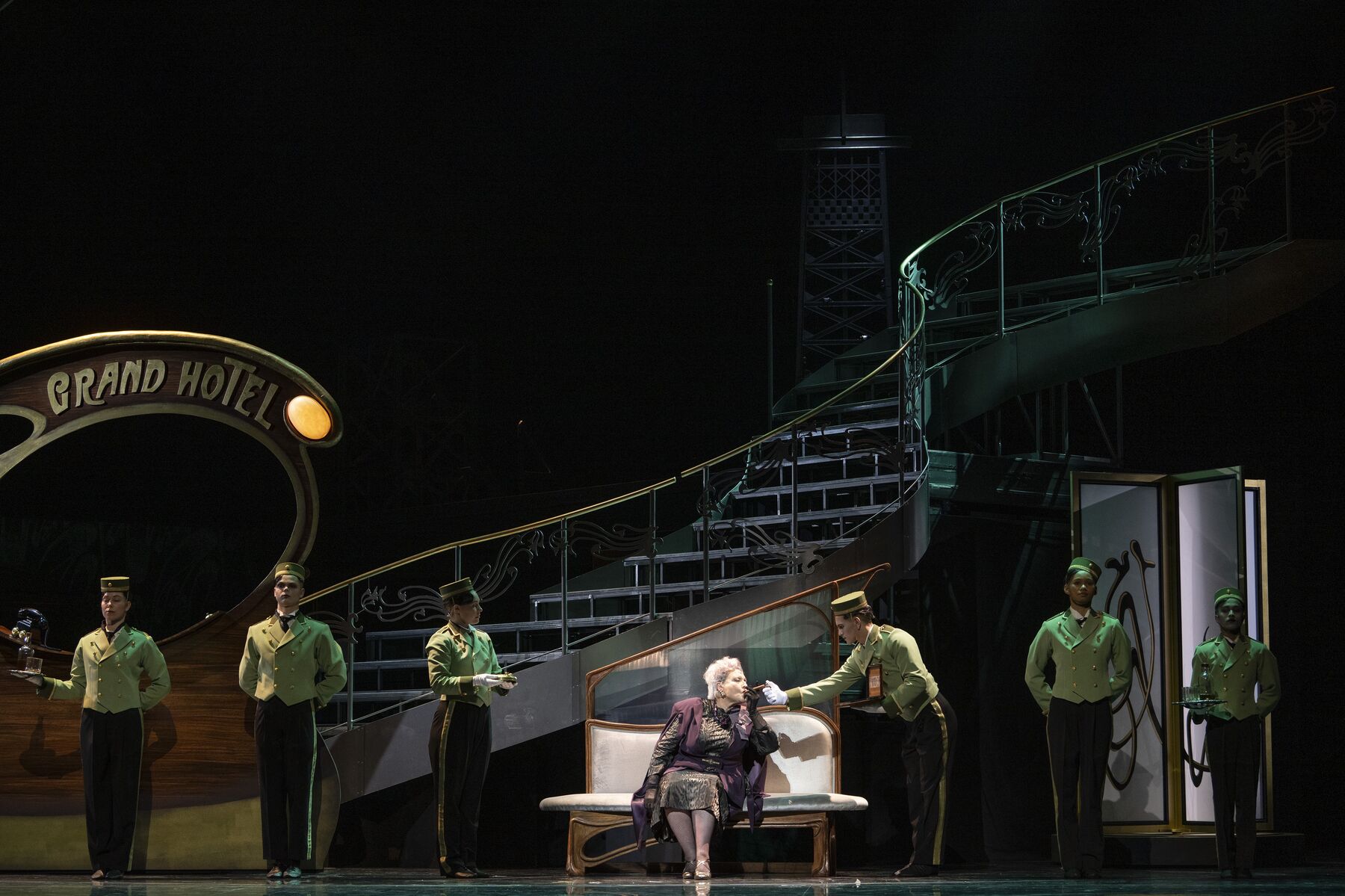 Eine ältere, schick gekleidete Frau sitzt auf einer weißen Bank in einer Hotellobby. Neben ihr stehen sechs Pagen in grüner Uniform. Einer von ihnen zündet der Frau ihre Zigarre an.