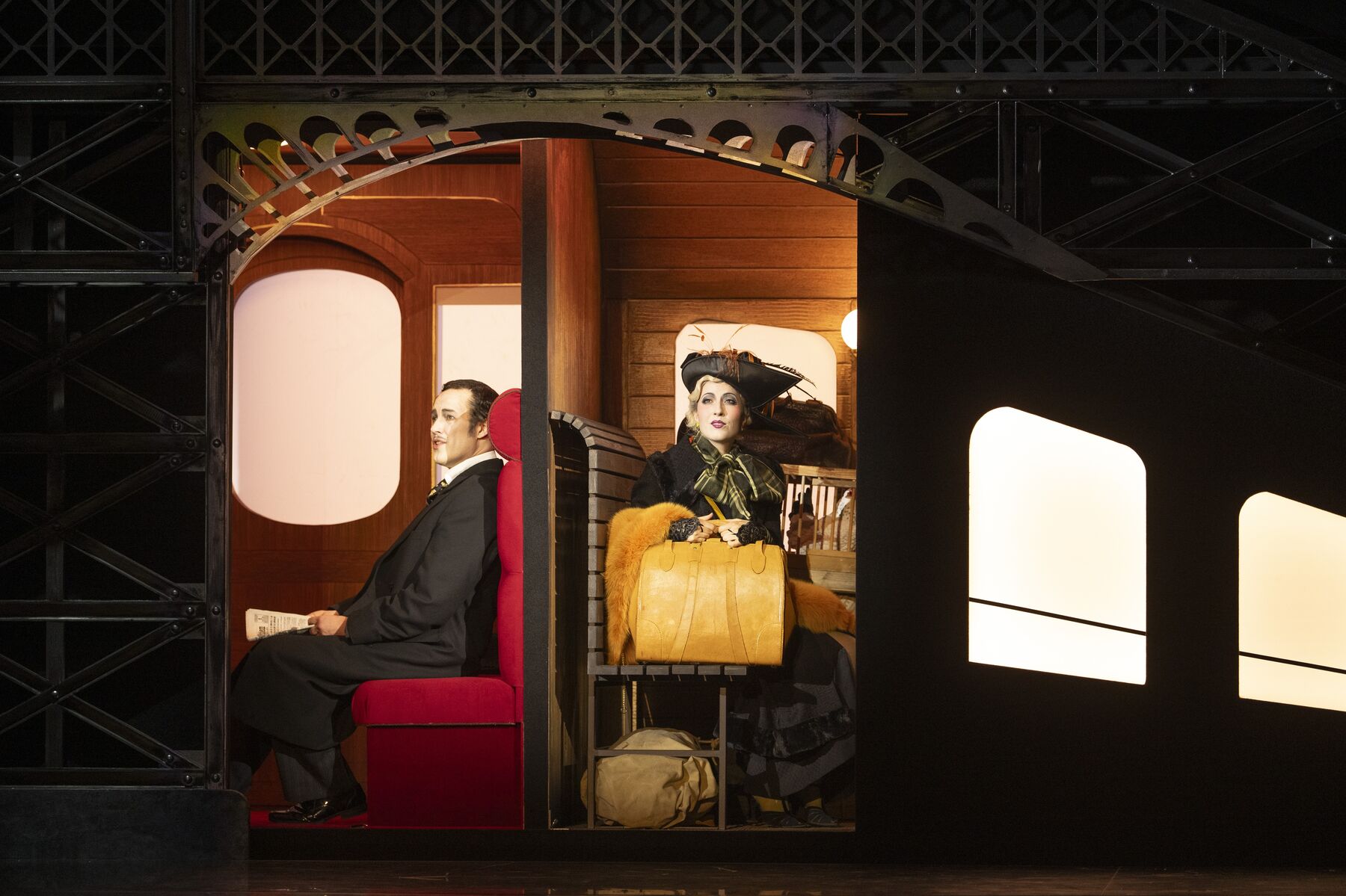 ein elegant gekleideter Mann in einem Anzug sitzt in einem schönen Zugabteil. Recht neben ihm sitzt eine pompös gekleidete Frau in einem 3. Klasse Abteil.