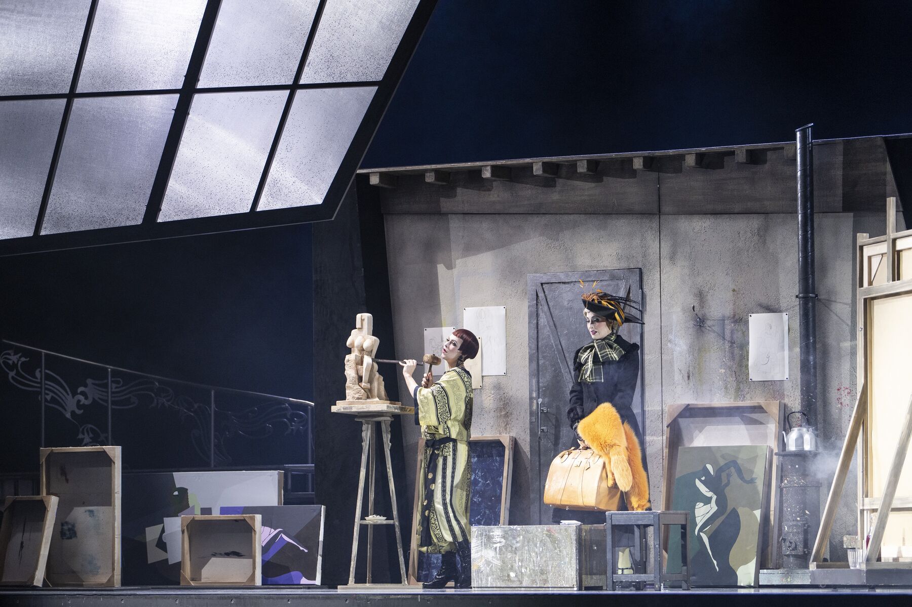 Zwei Frauen stehen in einem dunklen Atelier mit vielen Leinwänden. Die linke Frau arbeitet an einer Skulptur. Die rechte Frau steht hinter ihr und hält eine Tasche und einen Fellschal in ihrer Hand.