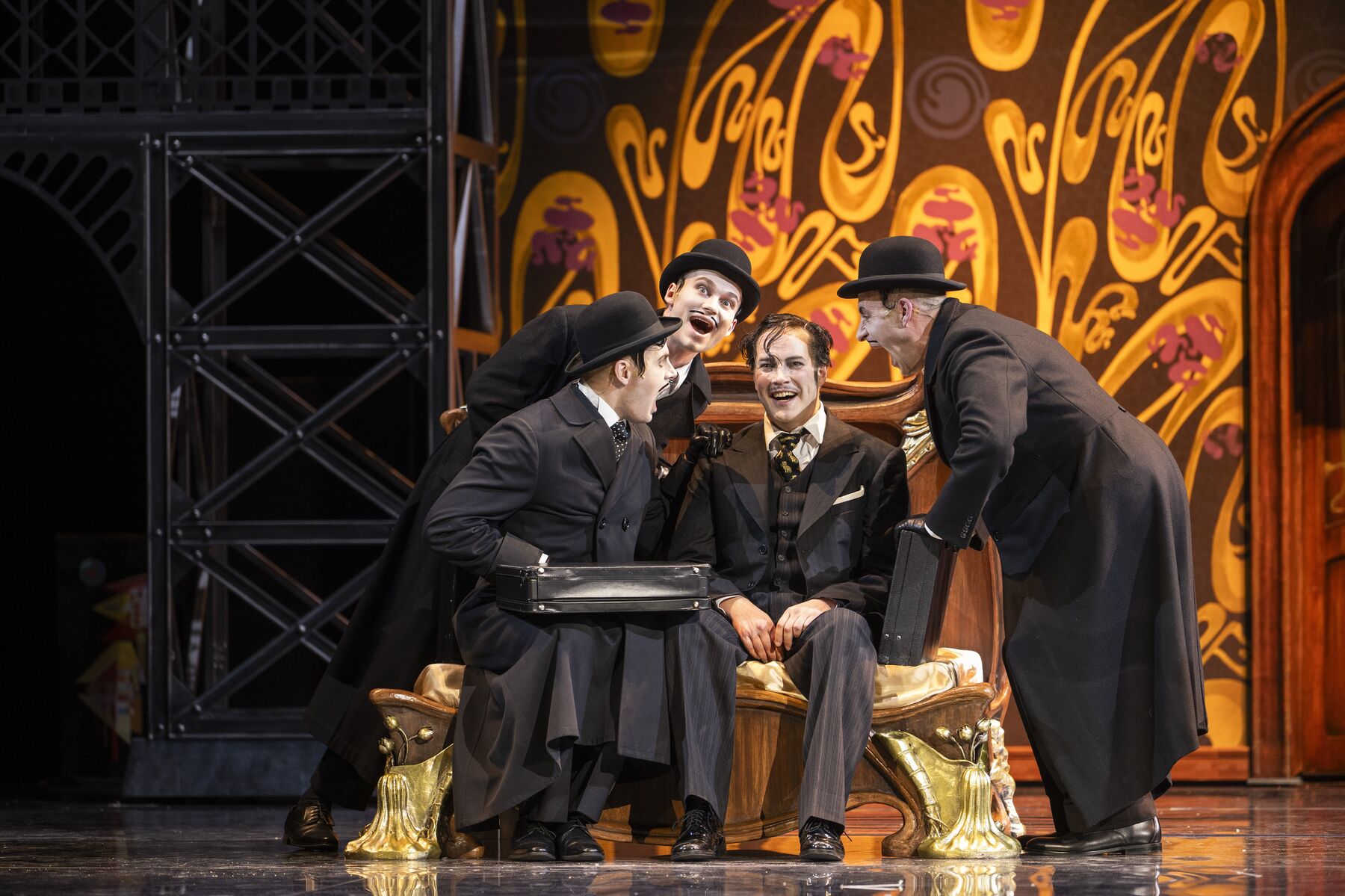 ein Mann in einem schwarzen Anzug sitzt an der Kante eines Betts. Um ihn herum stehen drei Männer in schwarzen Mänteln und mit schwarzen Hüten, die ihn anschauen.