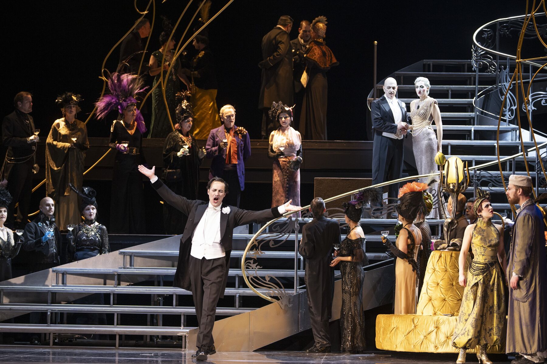 Ein Mann in einem schwarzen Frack steht singend mit offenen Armen vor einer langen Treppe und einer festlich gekleideten Menschenmasse.
