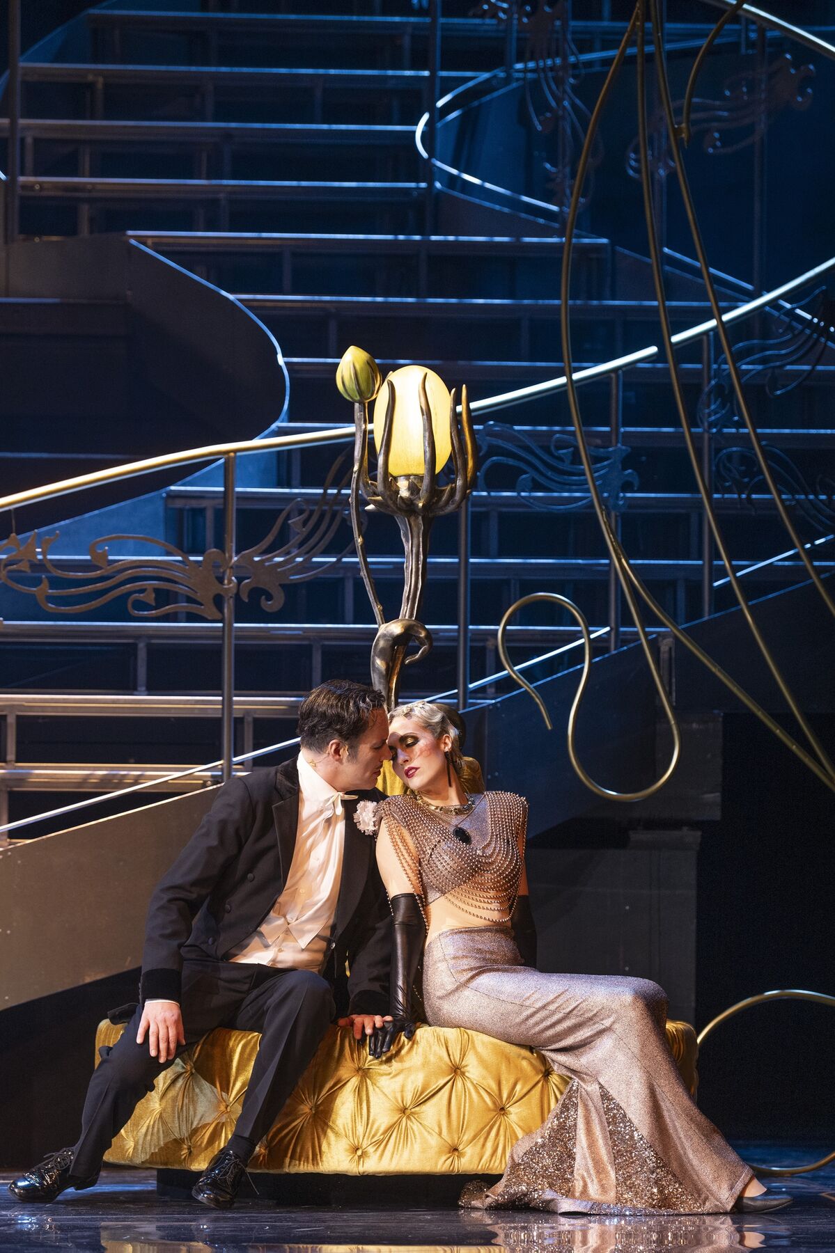 eine Mann in einem schwarzen Frack und eine Frau in einem engen silbernen Kleid sitzen auf einem schönen, gelben Sofa und nähern sich einem Kuss an.