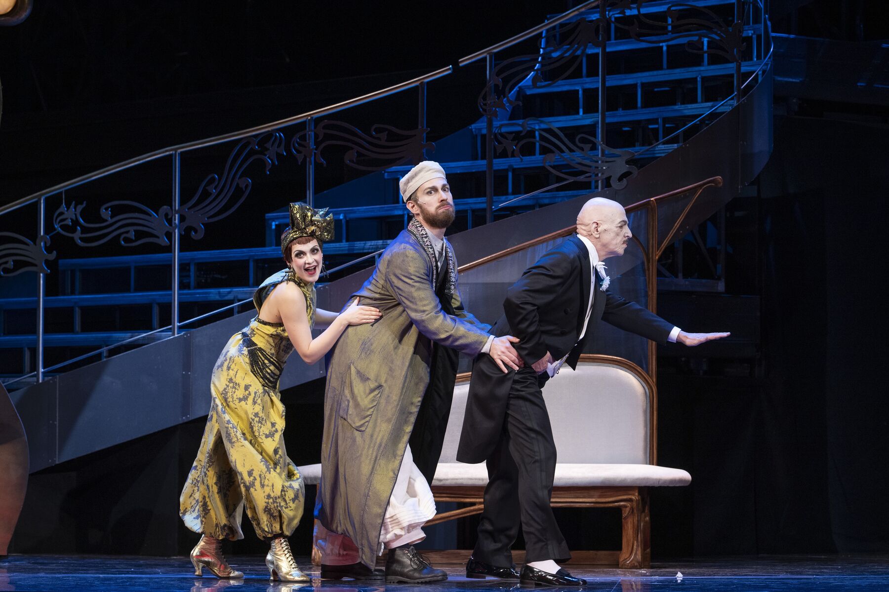 Eine Frau in einem gelben Hosenanzug, eine Mann in einem orientalisch angehauchten Kostüm und ein Mann in einem schwarzen Frack, stehen hintereinander und halten sich am jeweils vorderen fest.