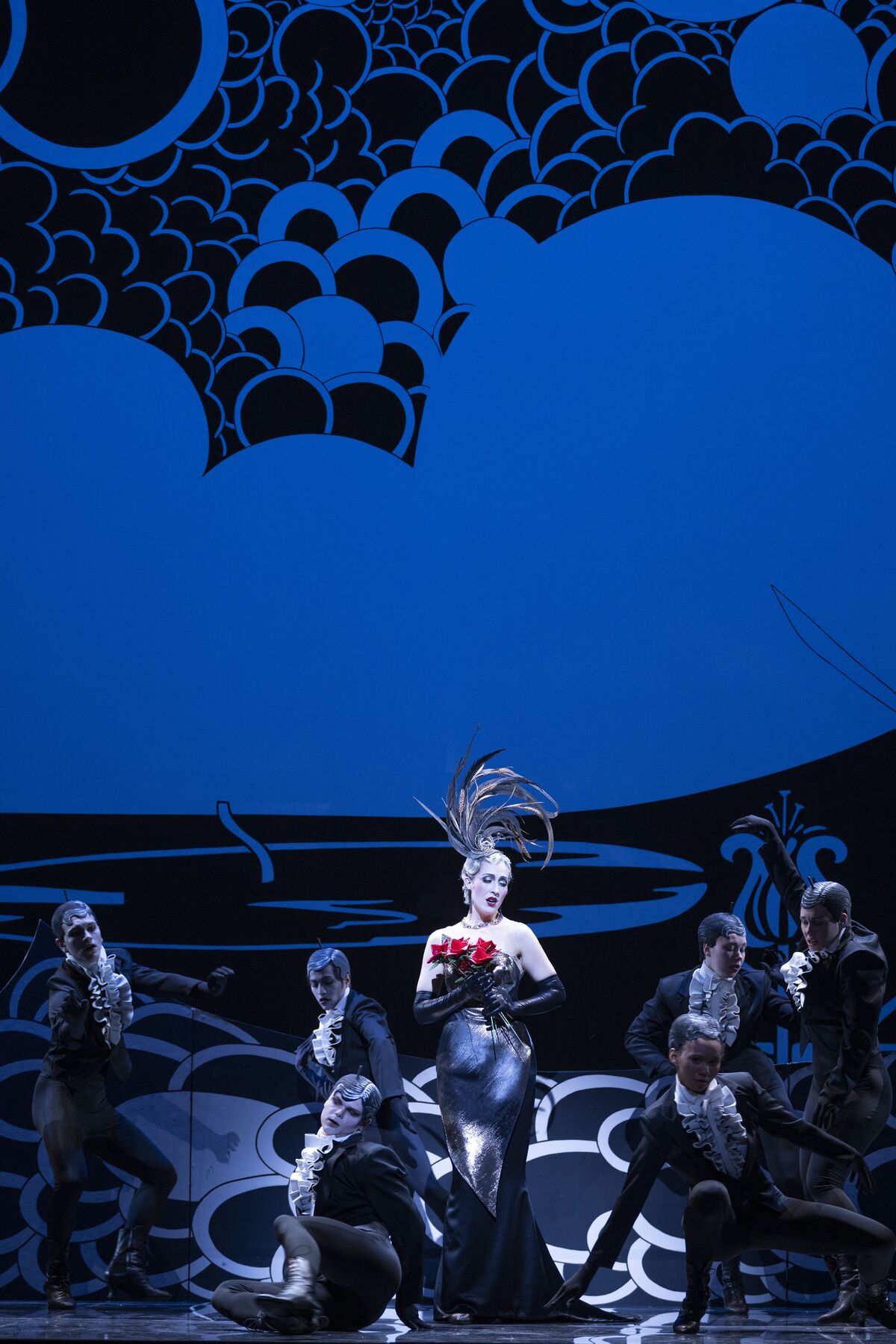 eine Frau in einem engen schwarzen Kleid und einer Kopfbedeckung mit Federn steht mit Blumen in der Hand zwischen schwarzgekleideten Tänzern.