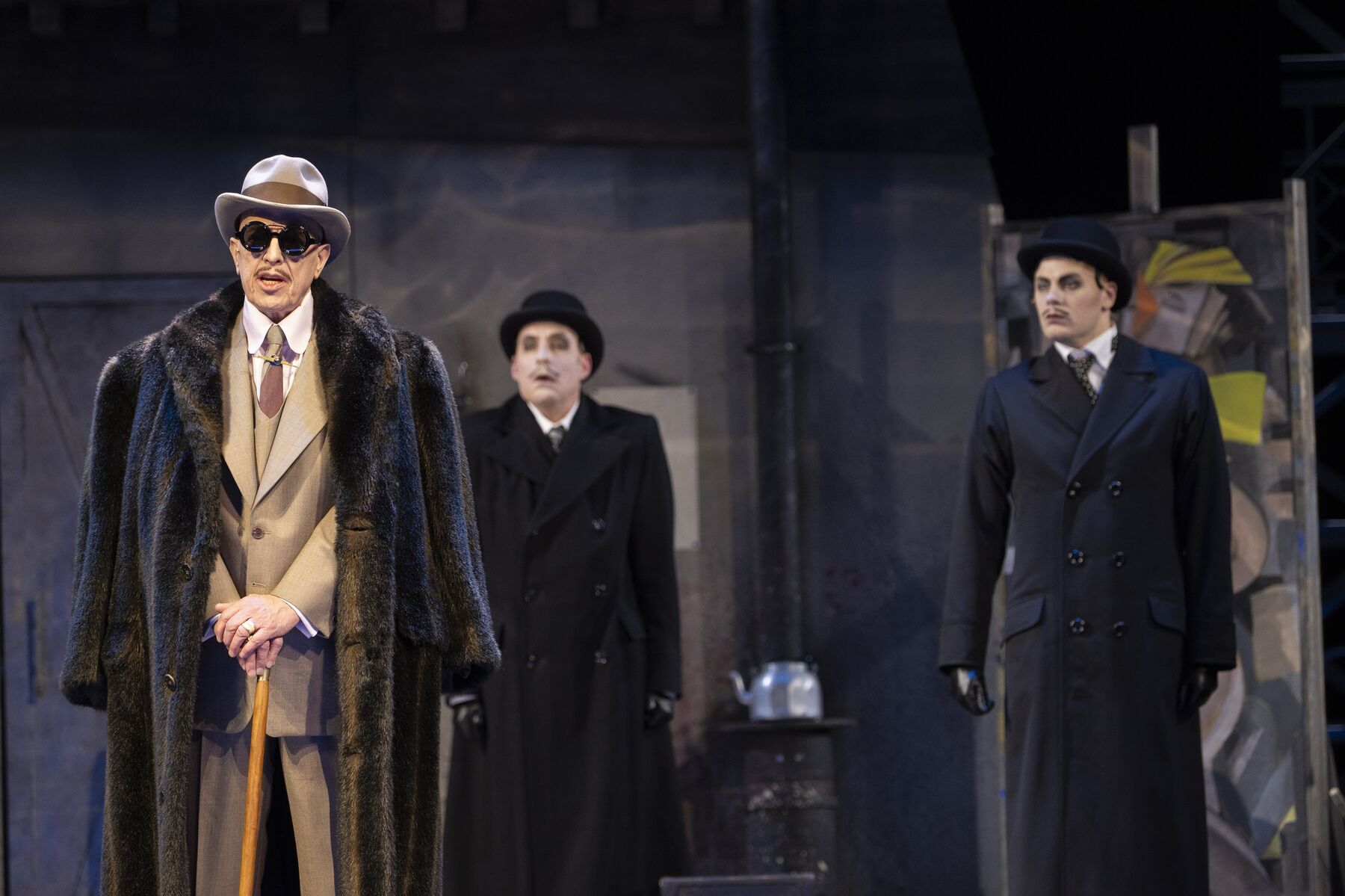 Ein Mann in einem hellbraunen Anzug und ´Fellmantel steht in einem dunklen Raum. Links hinter ihm stehen zwei Männer in langen schwarzen Mänteln und mit schwarzen Hüten.