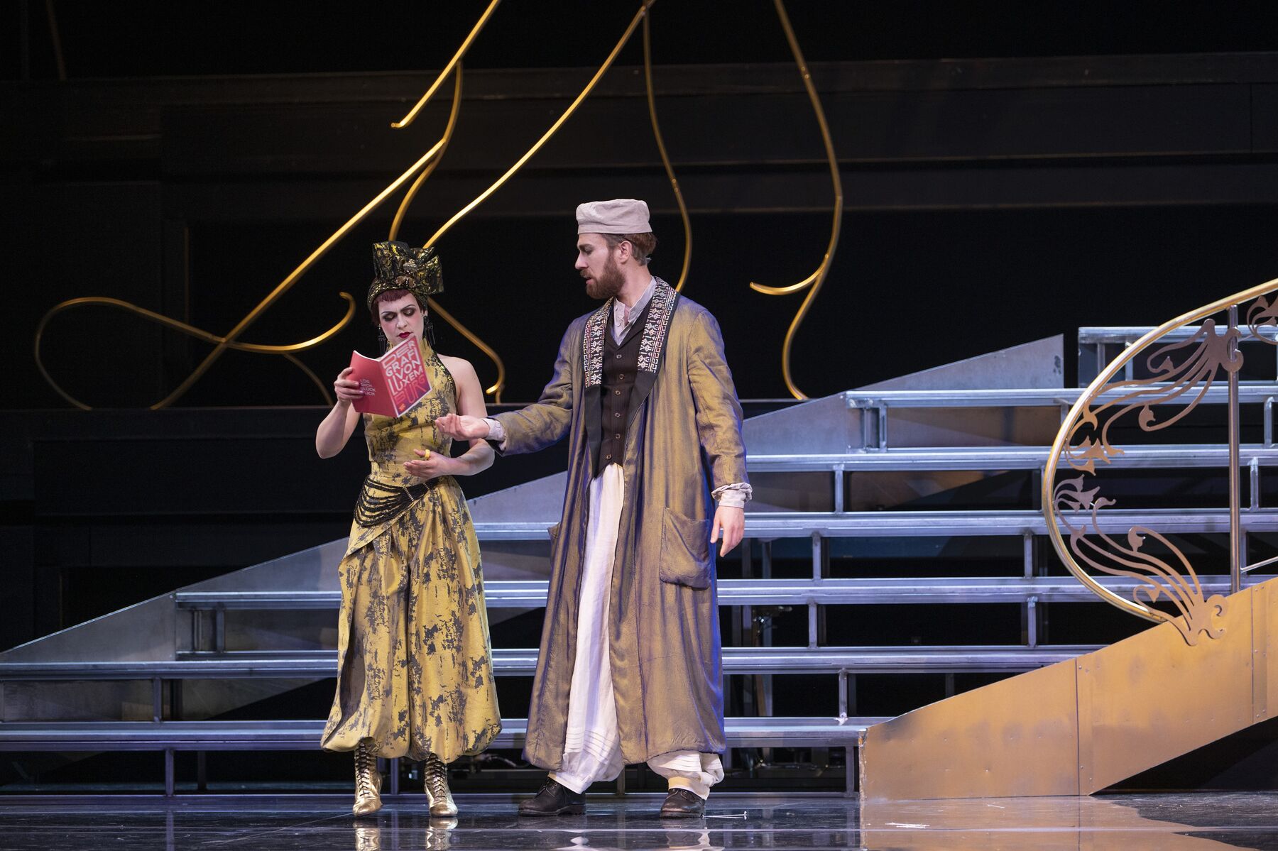 eine Frau in einem kunstvollen gelben Hosenanzug hält ein pinkes Programmheft in der Hand. Links neben ihr steht ein Mann mit einem orientalisch angehauchten Kostüm, der nach dem Heft greift.