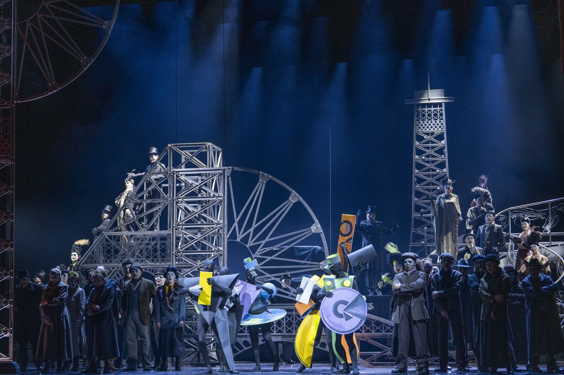 Tänzer in geometrischen Kostümen stehen zwischen einer Menschenmasse in dunkler Kleidung. Im Hintergrund ist der Eifelturm, weitere Gebäude in Stahloptik und eine Treppe zusehen.