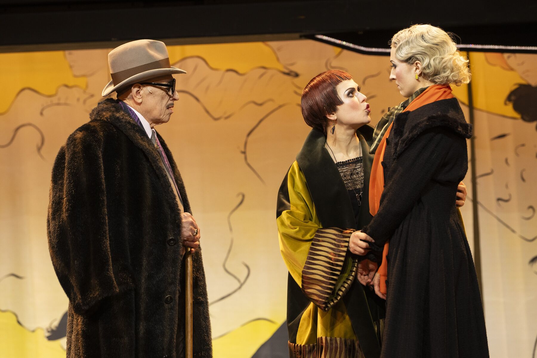 Ein Mann mit einem Hut und einem braunen Fellmantel schaut zwei Frauen an. Die eine Frau steht leicht erschrocken da und schaut die Andere an, welche sie mahnend anschaut.