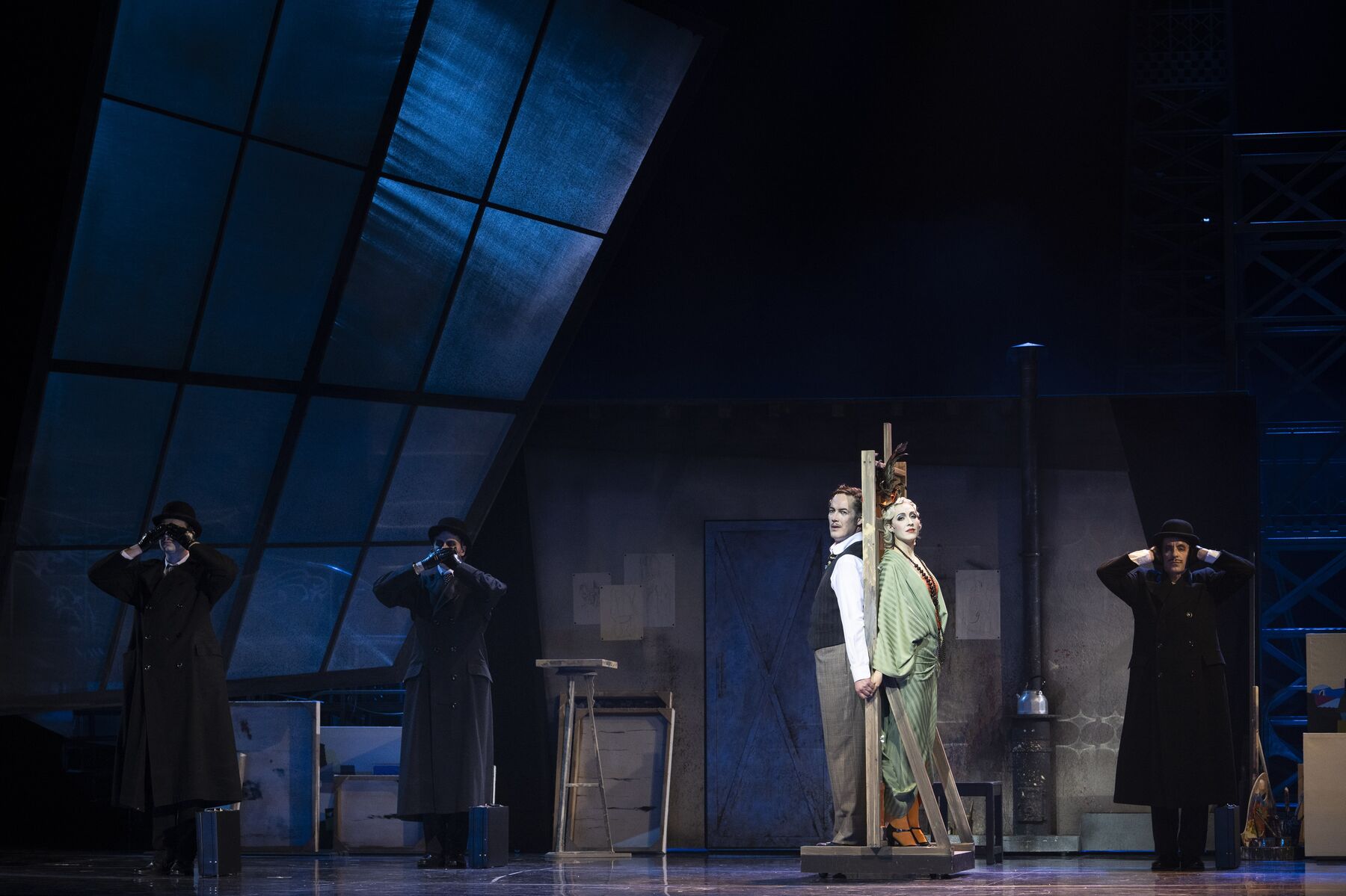 Ein Mann und eine Frau stehen mit dem Rücken zueinander und durch eine dünne Wand getrennt, in einem dunklen Raum. Um sie herum stehen drei Männer in schwarzen Mänteln und schwarzen Hüten. Sie halten sich entweder den Mund, die Augen oder die Ohren zu.