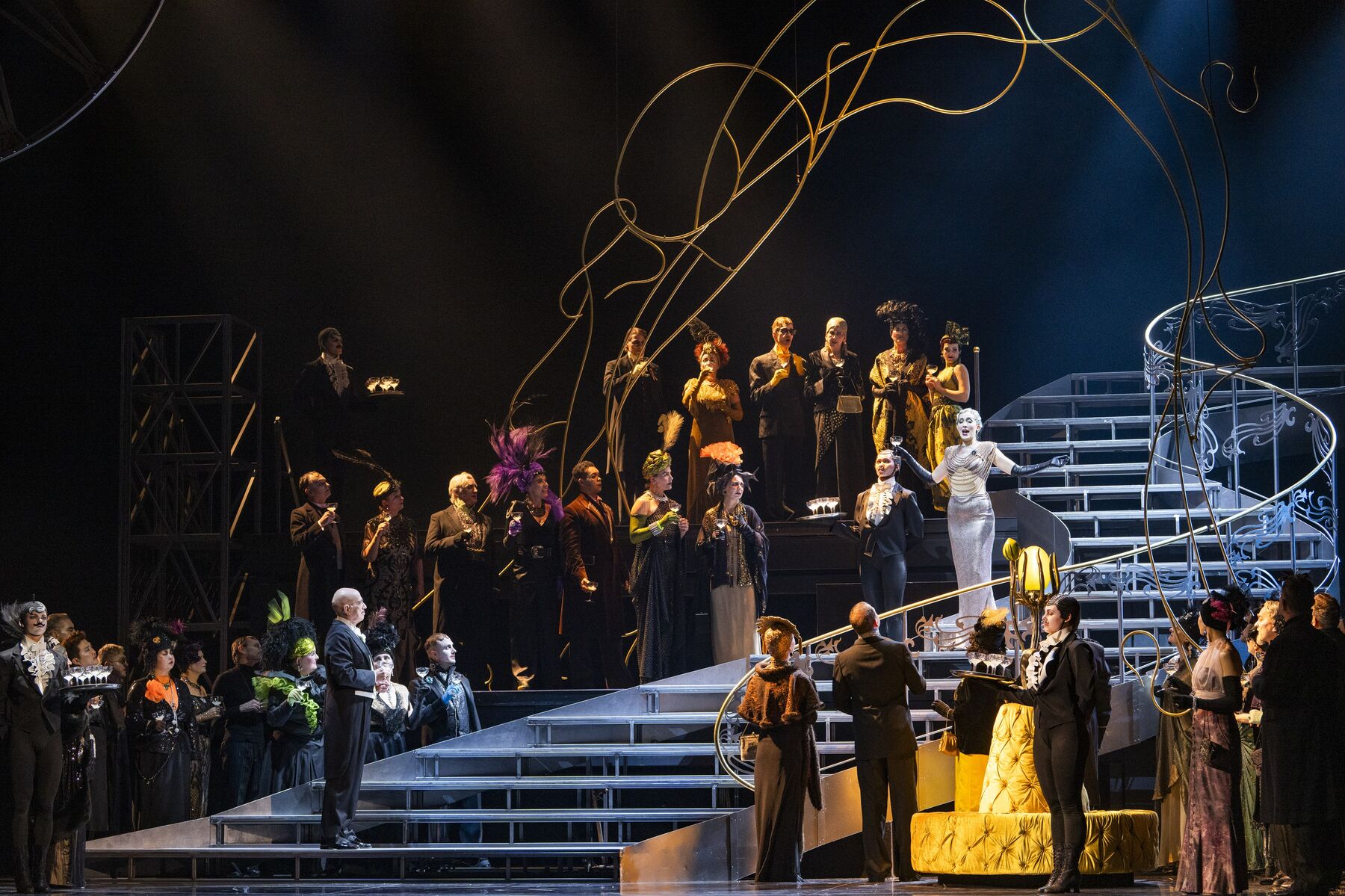 Festlich gekleidete Menschen stehen um eine Lange Treppe. Auf der Treppe steht eine Frau in einem engen silbernen Kleid. am Ende der Treppe steht ein Mann mit einem schwarzen Anzug der sie anschaut.