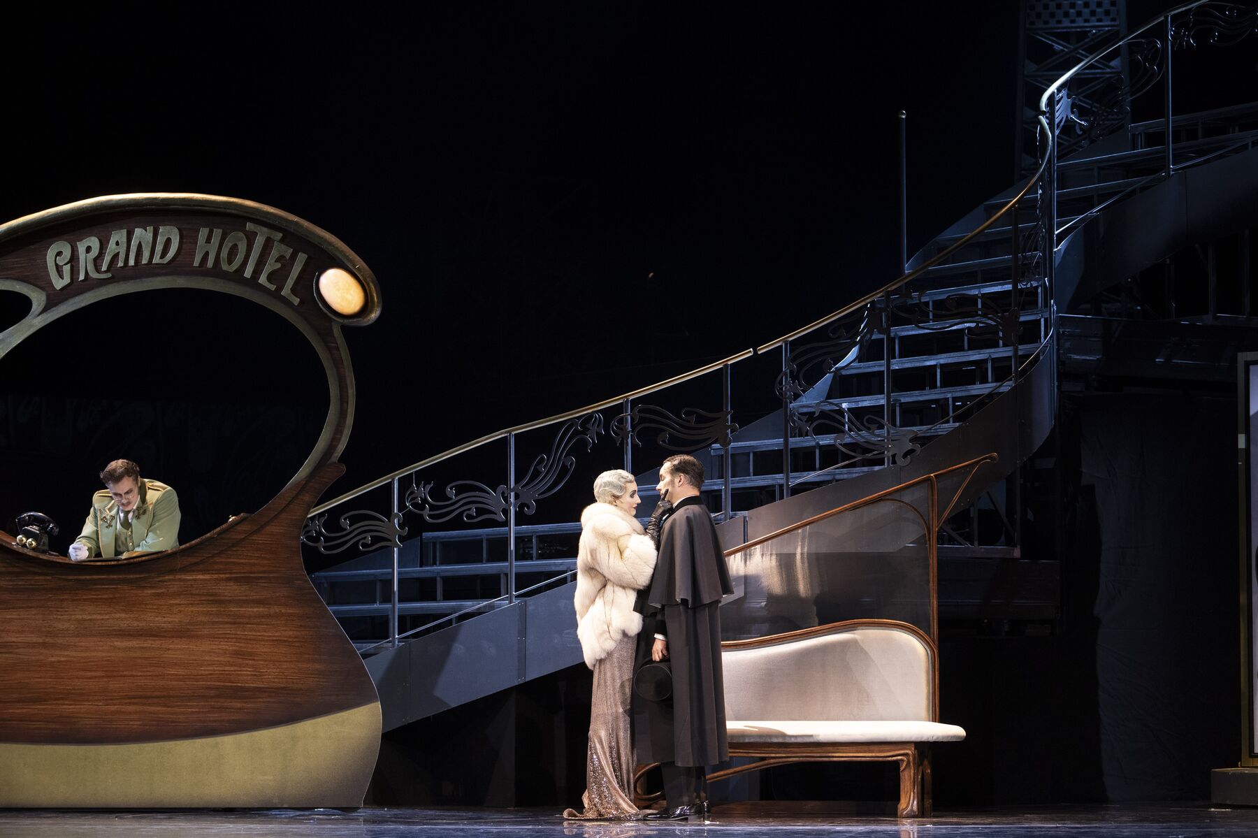 eine Frau mit einem silbernen Kleid und weißem Fellmantel und ein Mann in einem eleganten schwarzen Mantel stehen eng beieinander in einer Hotellobby. Links hinter ihnen ist die Rezeption mit einem Portier.