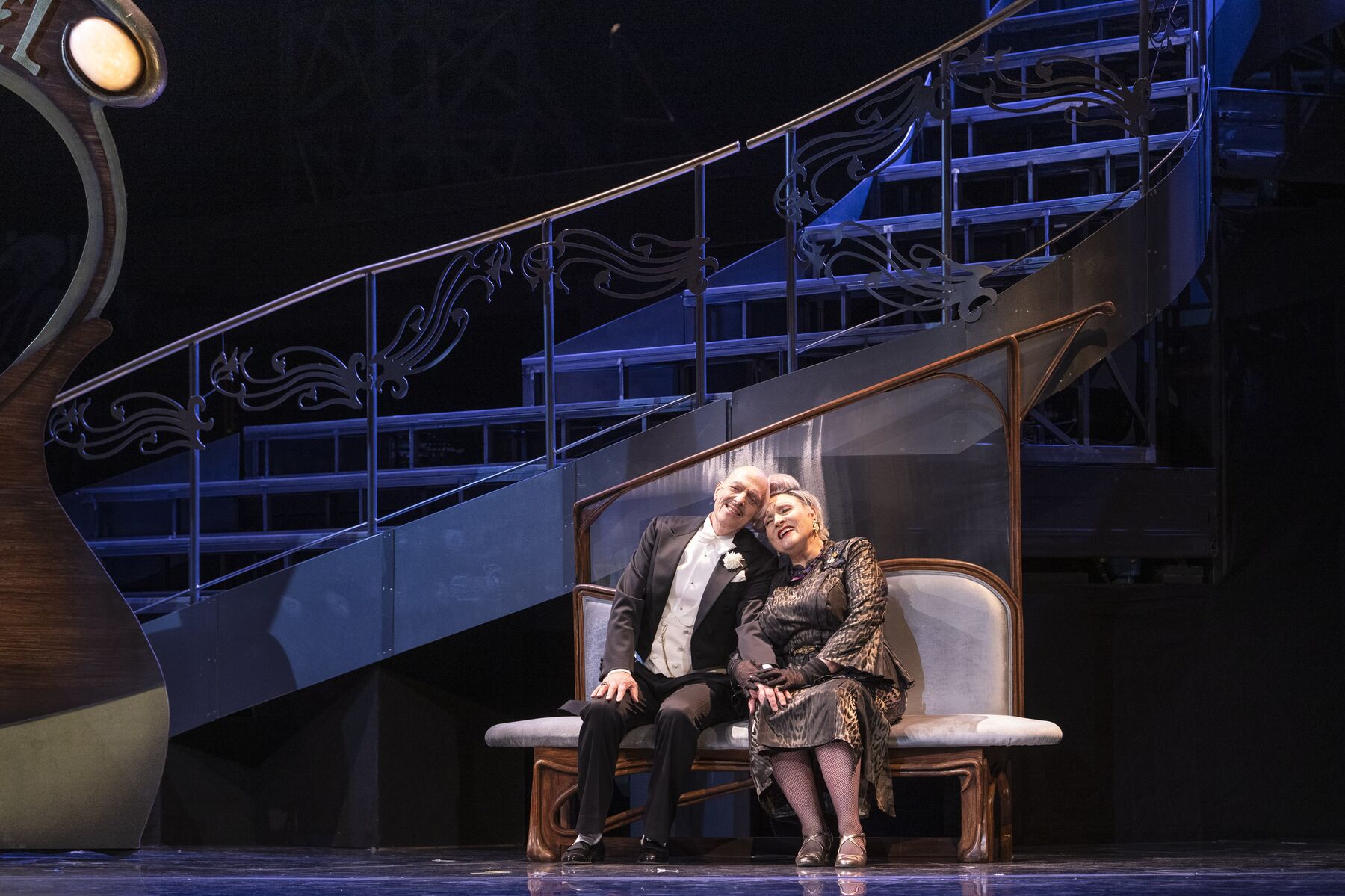 eine elegant gekleidete ältere Dame und ein älterer Mann in einem Anzug sitzen auf einer weißen Bank. Sie lehnen ihre Köpfe liebend gegeneinander.