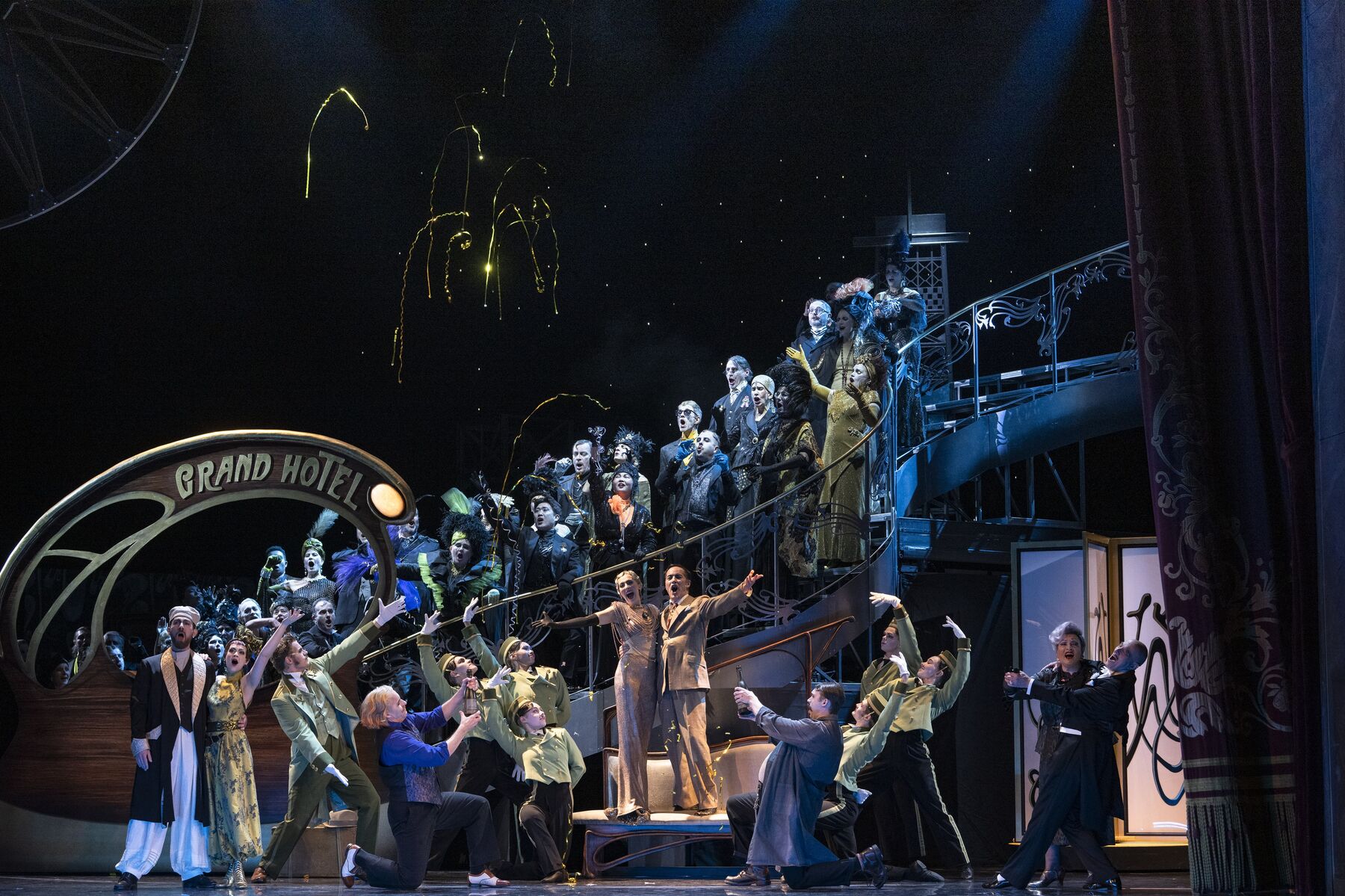 eine festlich gekleidete Menschenmenge steht auf einer Treppe und um eine weiße Bank. Auf der Bank steht ein Pärchen. Alle schauen freudig und jubelnd zu ihnen.