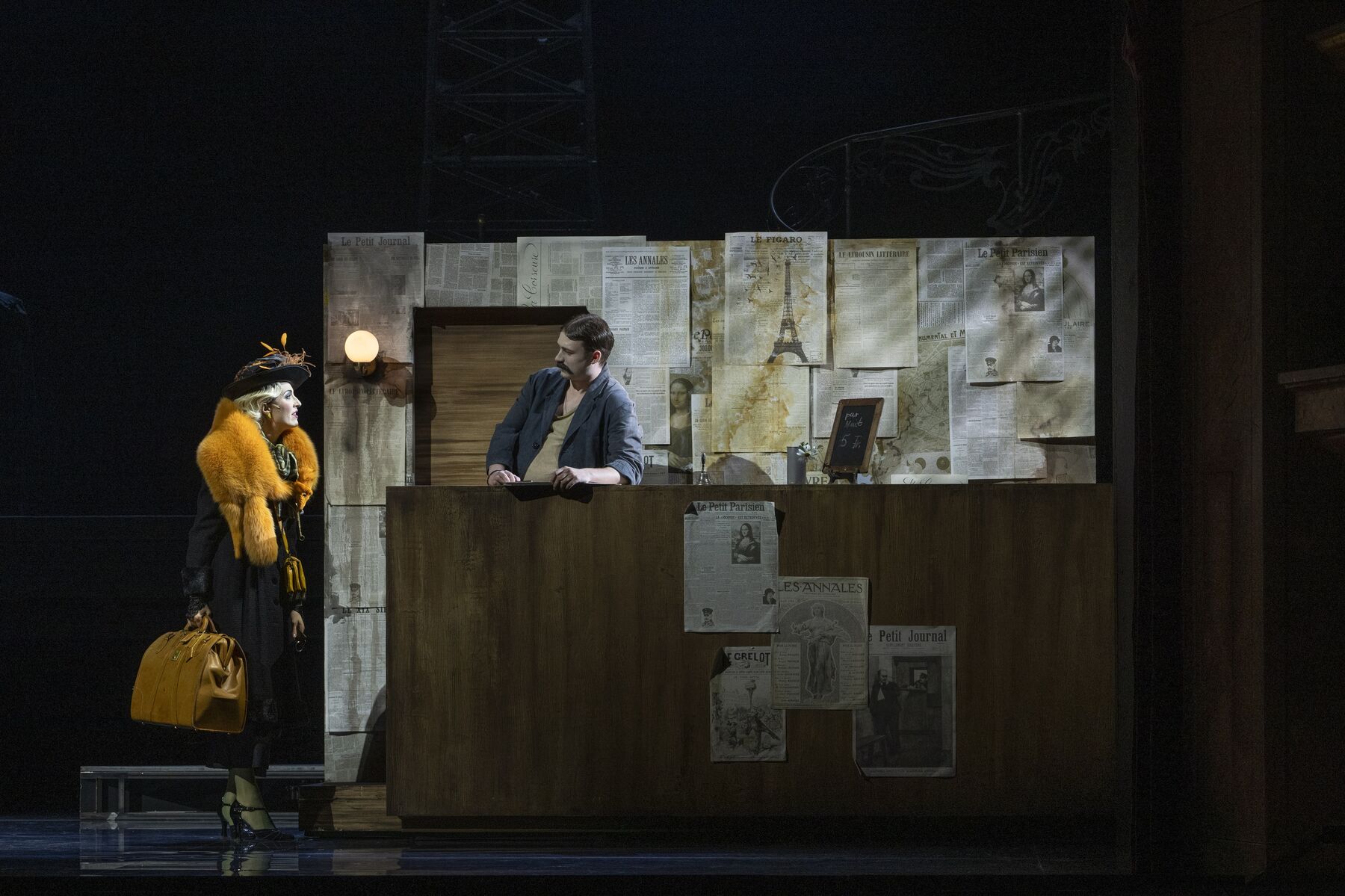 Ein Mann steht an einem Tresen. Hinter ihm ist eine Wand die mit Zeitungen tapeziert ist. Links neben ihm steht eine Frau mit einer großen Tasche und einem pompösen, gelben Fellkragen.