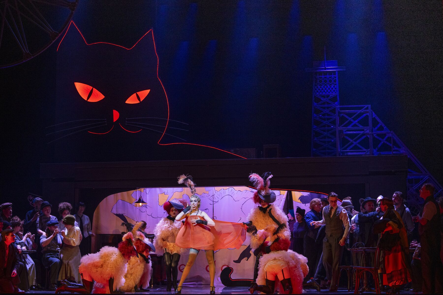 Eine Menschenmasse steht links und recht von einer Gruppe Tänzerinnen. Die Tänzerin in der Mitte ist leichtbekleidet. Die anderen Tänzer halten ein Tuch vor ihren Körper. Sie haben pompöse Hüte und Jacken an. Im Hintergrund ist in rotem Licht die Umrandung einer Katze zusehen.