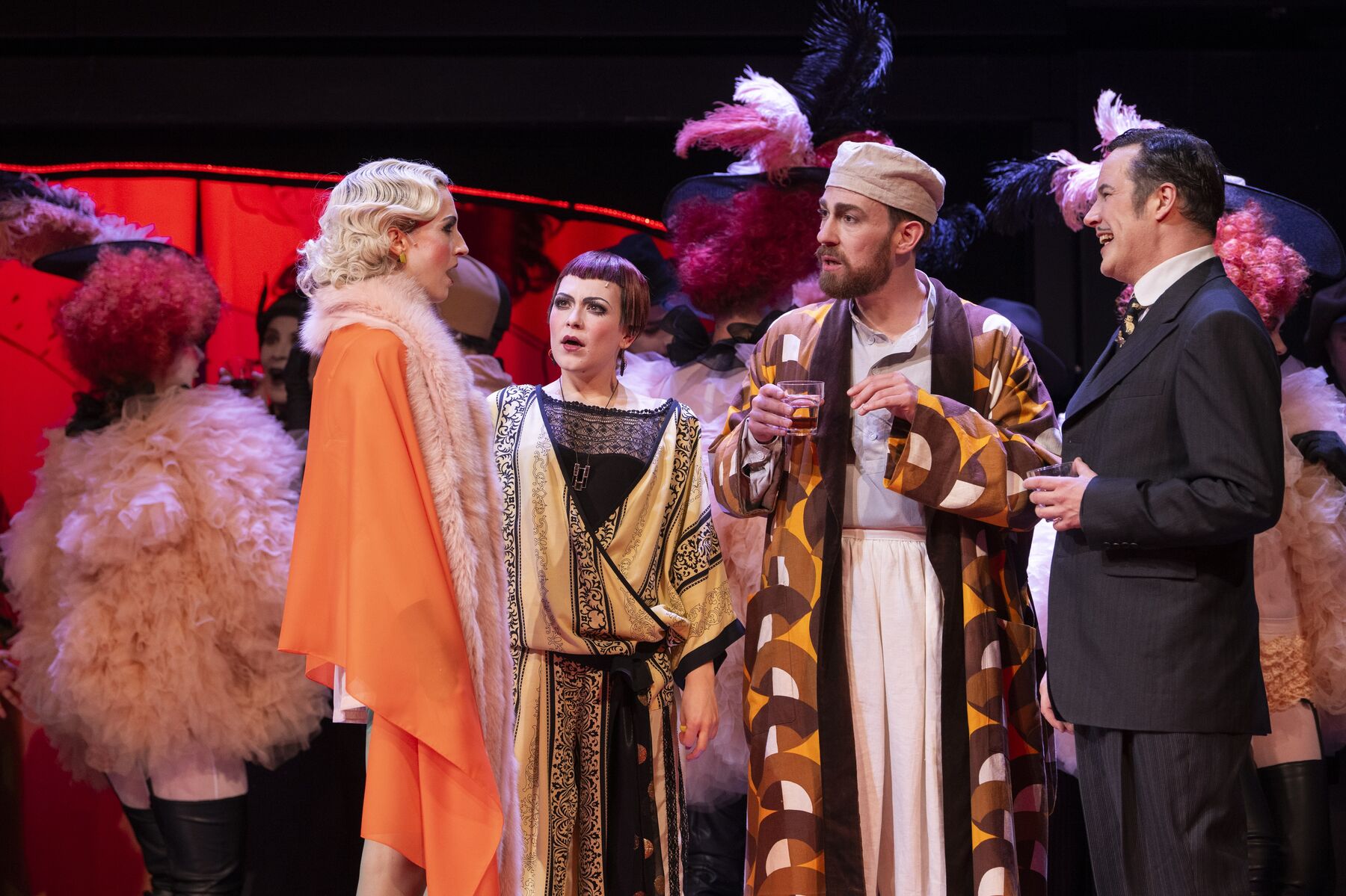eine Gruppe aus zwei Frauen und zwei Männern in festlicher Kleidung, die am Jugendstil orientiert ist reden miteinander. Im Hintergrund sind Tänzer in flauschigen Jacken zusehen.