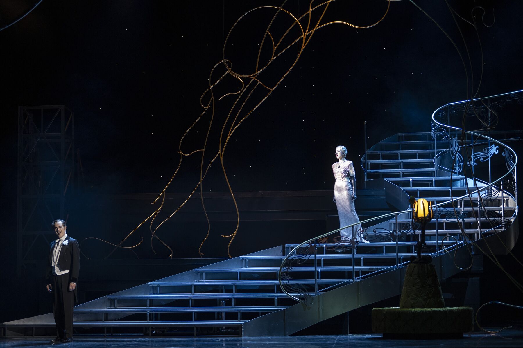 eine Frau in einem engen silbernen Kleid steht in der Mitter eine langen schwungvollen Treppe. Unten an der Treppe steht ein Mann mit schwarzem Frack der sich ihr abwendet.