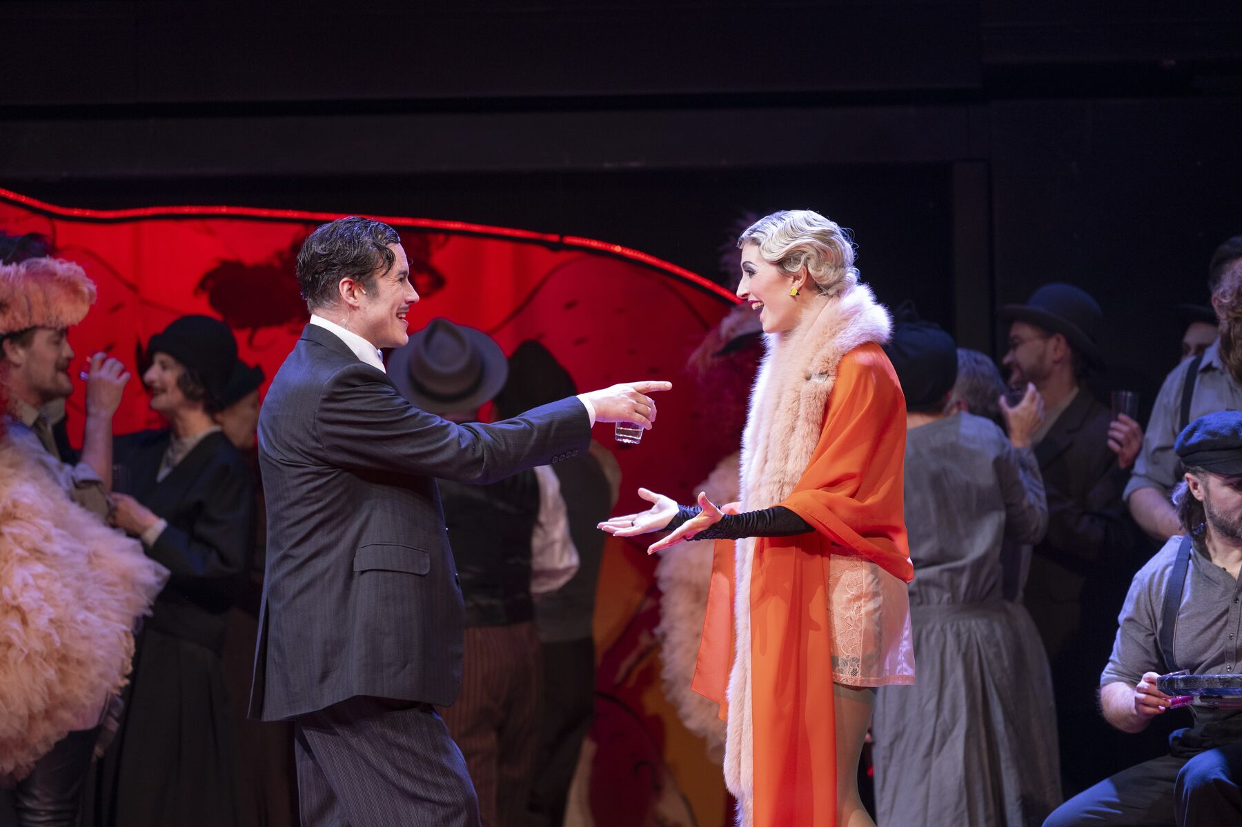 In der Mitte stehen ein Mann in einem schwarzen Anzug. Er zeigt fröhlich auf eine Frau die in einem roten Tuch und einem Fellschal gekleidet ist. Im Hintergrund sind weitere Menschen in festlicher Stimmung zusehen.