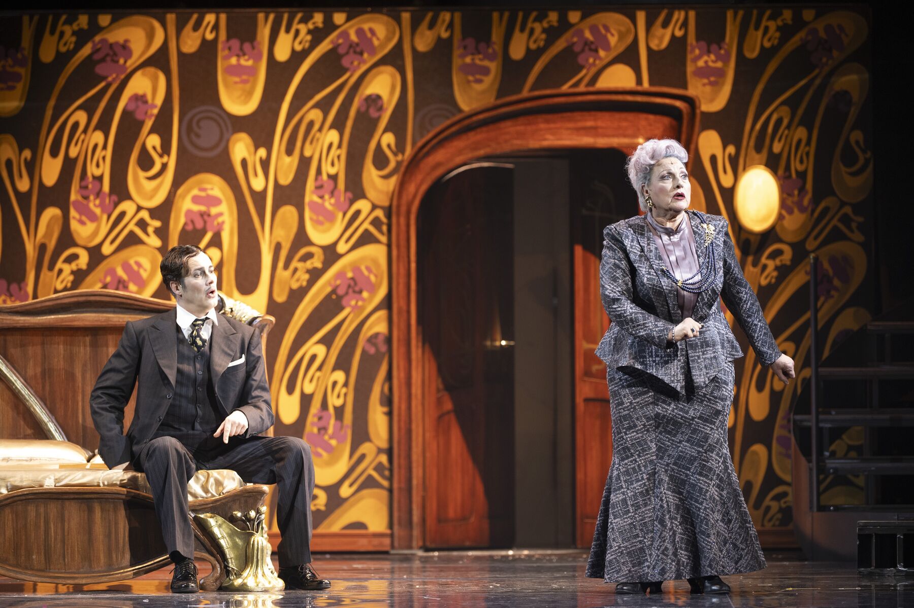 ein Mann in einem schwarzen Anzug sitzt auf einem Hotelbett. Er schaut eine älteren Dame hinterher die ein langen grauen Rock und einen grauen Blazer trägt.