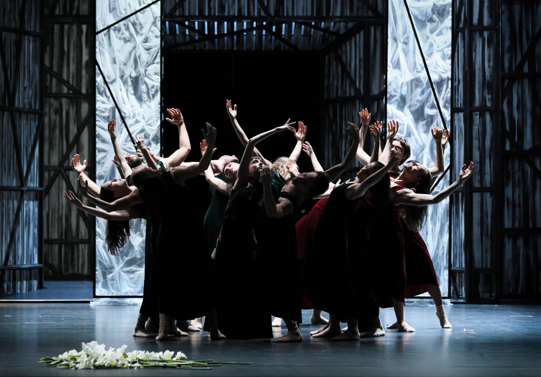 A group of ballet dancers stand together and lean back while stretching their hands in all directions. Their faces are brightly lit. In the background, you can see a black-and-white stage set that is partially illuminated. In the foreground, a bouquet of white flowers lies on the floor.