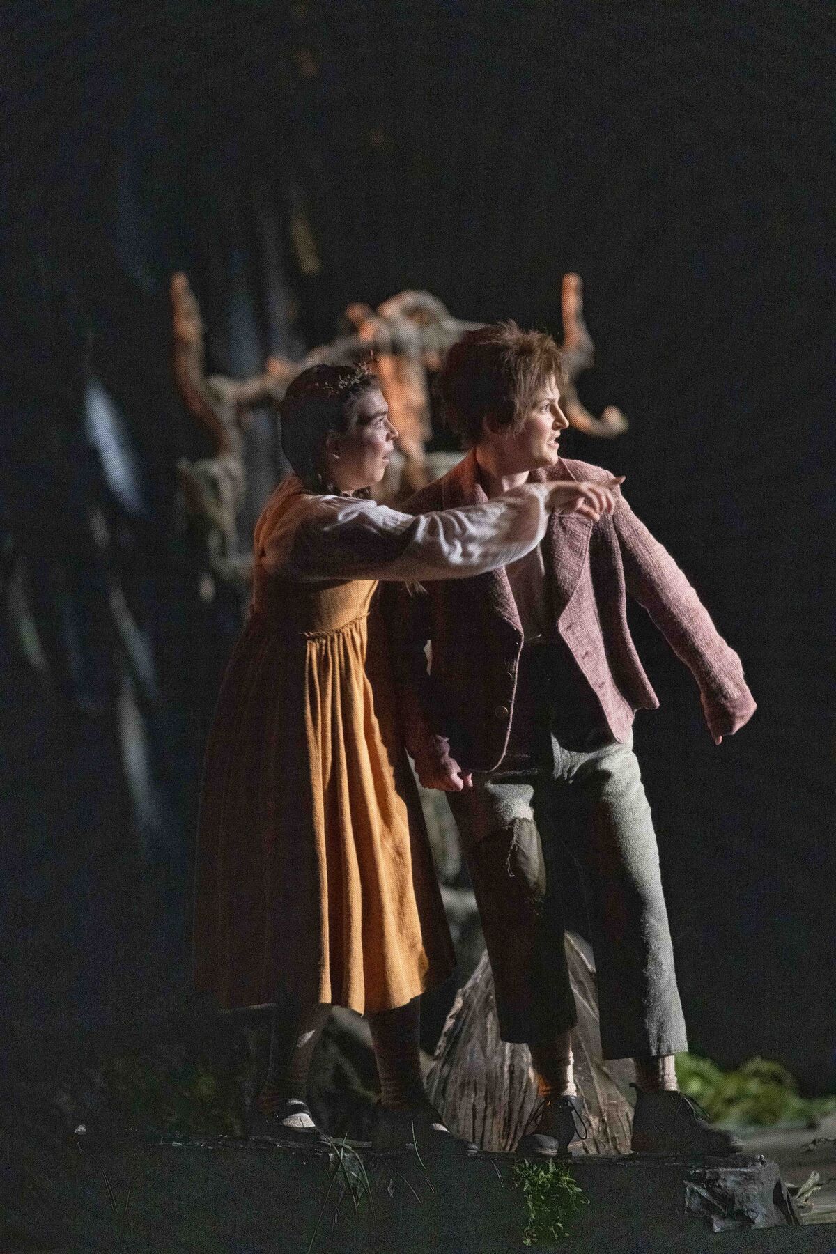 Das Bild zeigt die Darstellerinnen von Hänsel und Gretel von der rechten Seite. Beide sehen erschrocken aus, Gretel zeigt auf etwas. Die Szene ist sehr dunkel.