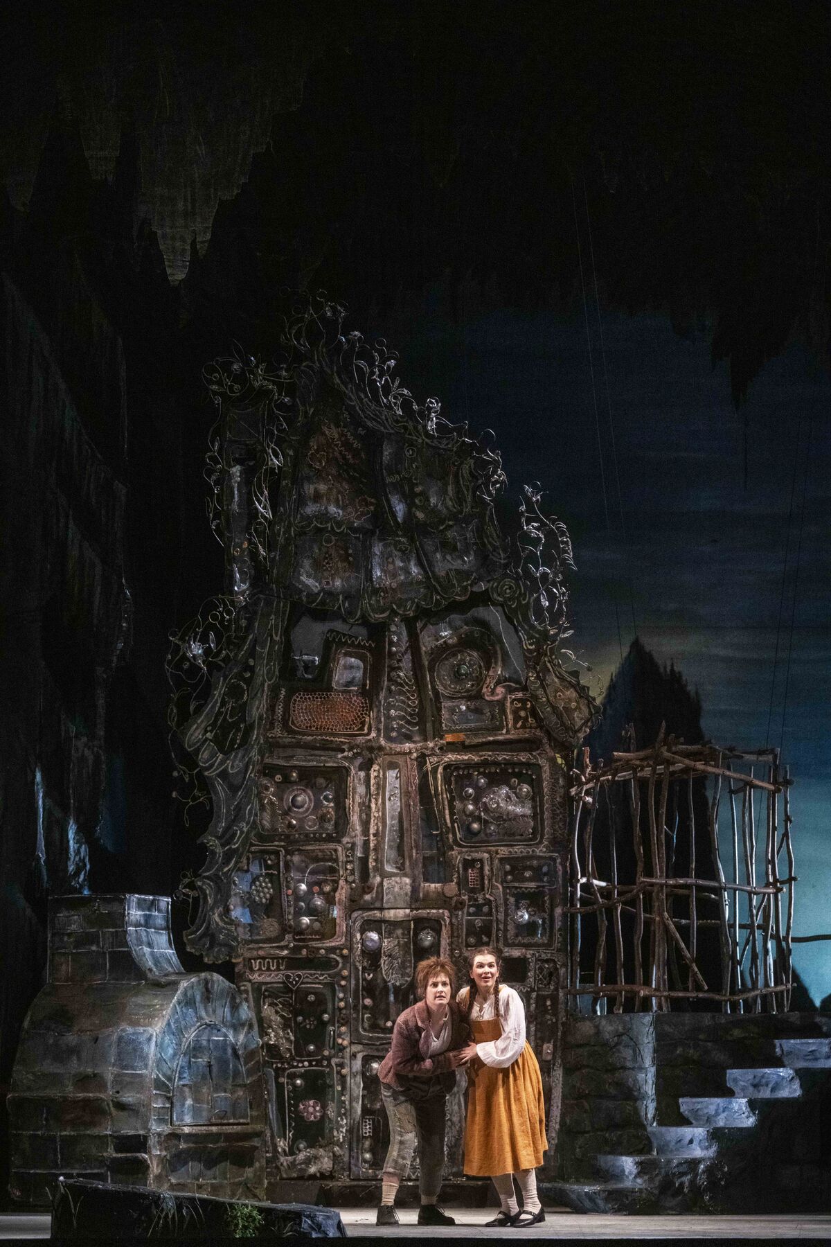 Hänsel und Gretel stehen überrascht vor dem Haus der Hexe. Das Haus ist düster und von Ranken umschlungen und mit Lebkuchen geschmückt.  Links im Bild steht ein steinerner Ofen, rechts ist ein hölzerner Käfig.