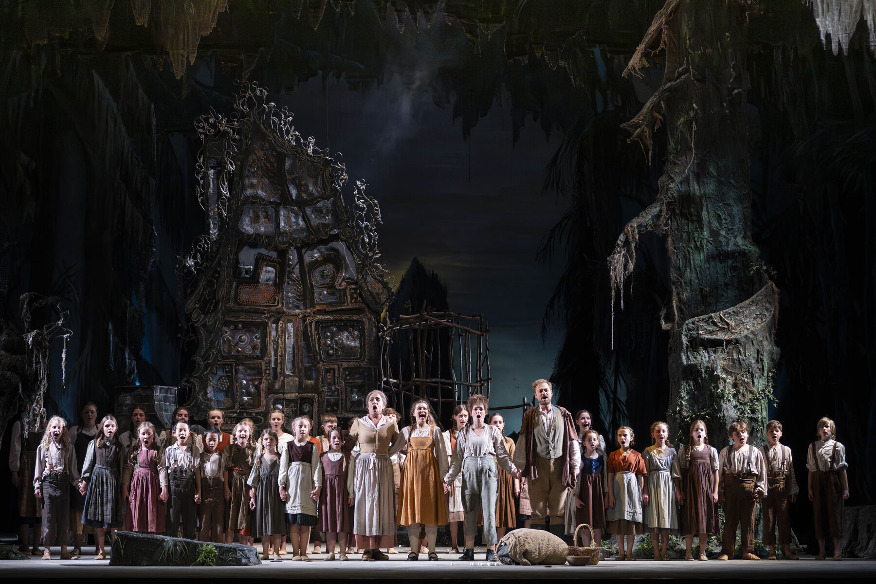 Hänsel, Gretel und ihre Eltern halten sich an den Händen. Sie sind wieder vereint. Hinter ihnen steht der Kinderchor. Sie singen vor dem Hexenhaus.