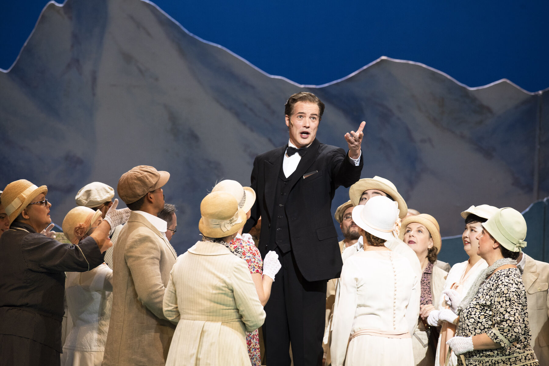 Ein Mann im schwarzen Anzug steht etwas erhöht und singt. Um ihn herum steht der Chor des Staatstheaters. Im Hintergrund sieht man Berge.