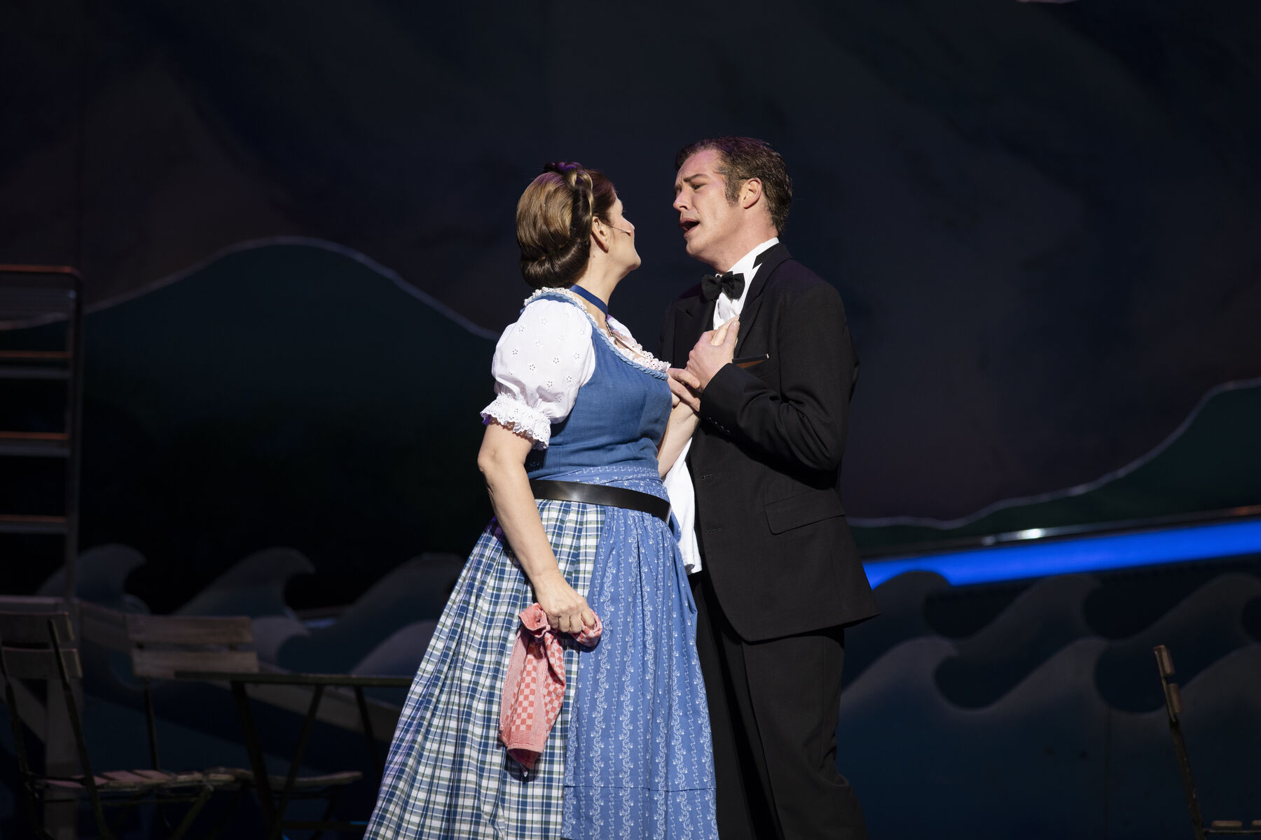 Eine Frau im blauen Dirndl steht vor einem Mann im schwarzen Anzug. Sie schauen sich an und singen.