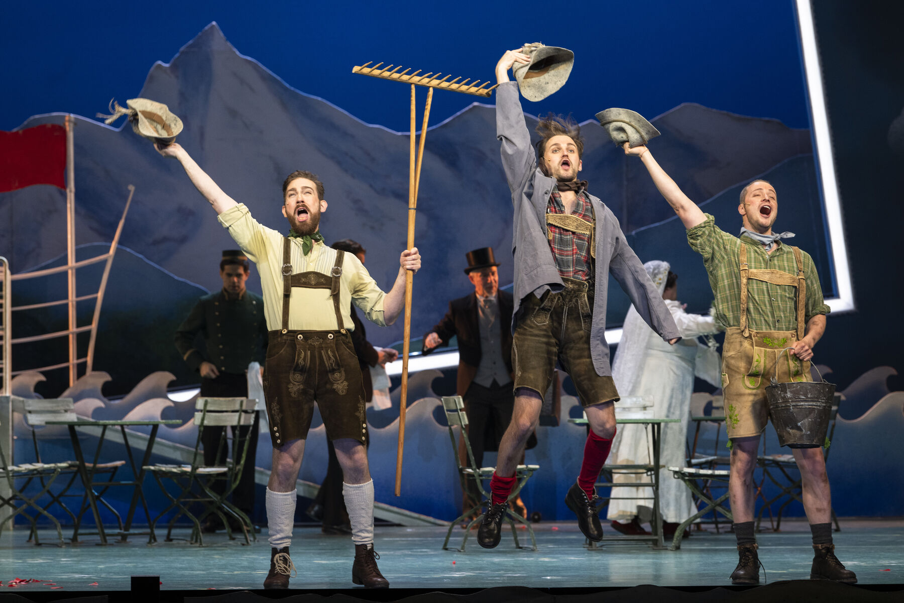Drei Männer in Tracht stehen und springen auf der Bühne. Im Hintergrund sieht man weitere Darstellerinnen.