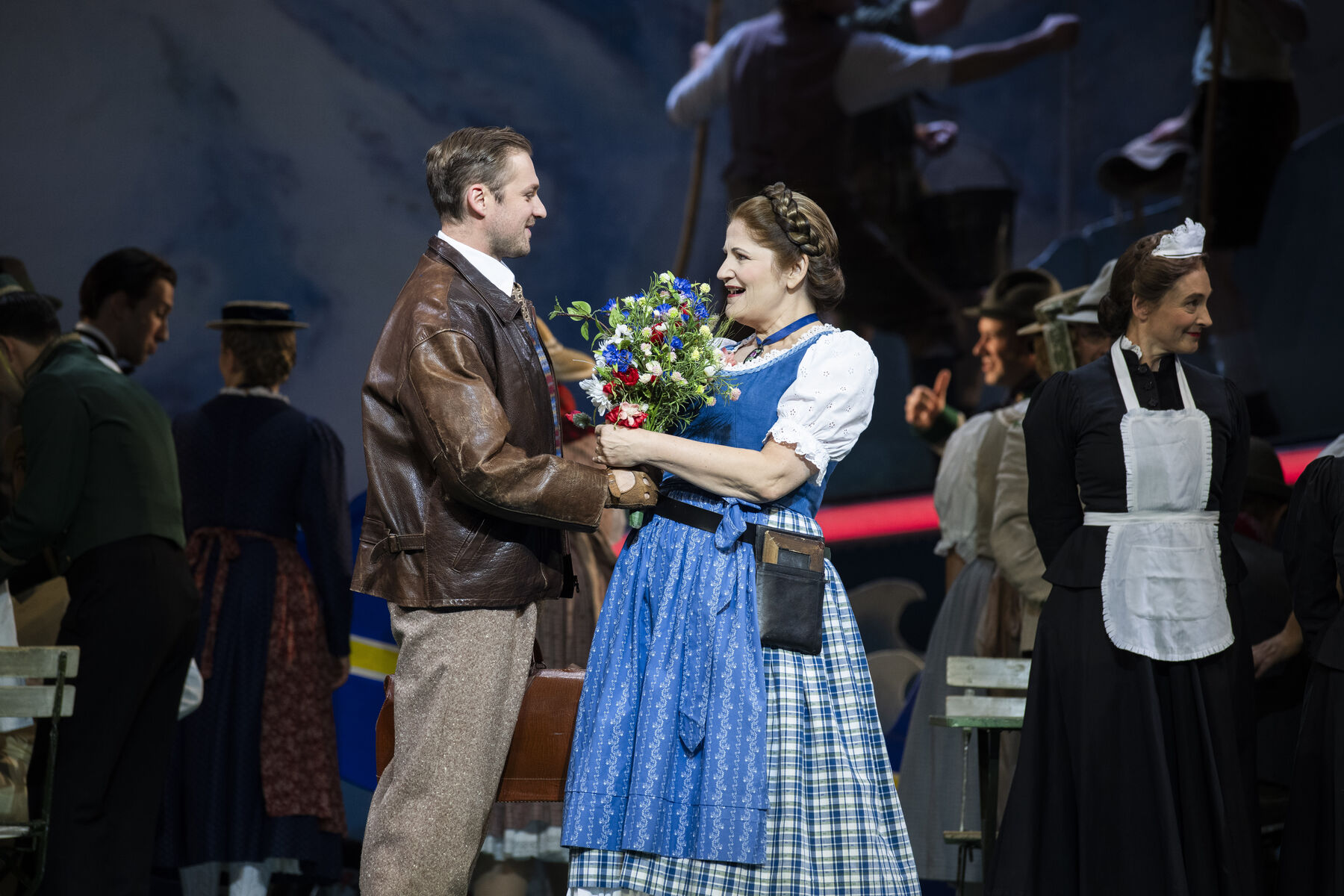 Ein Mann in Lederjacke übergibt einen Strauß Blumen an eine Frau im blauen Dirndl. Im Hintergrund sieht man weitere Darstellerinnen.
