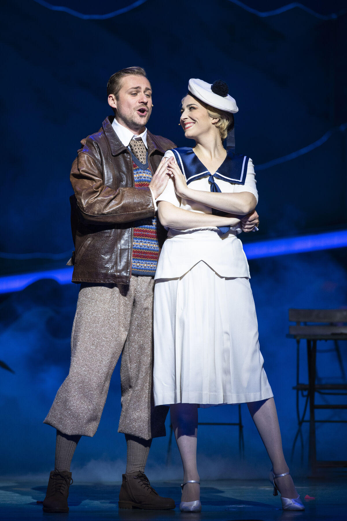 Ein mann in Lederjacke steht neben einer Frau im Matrosen-Anzug auf der Bühne. Die Bühne ist blau.