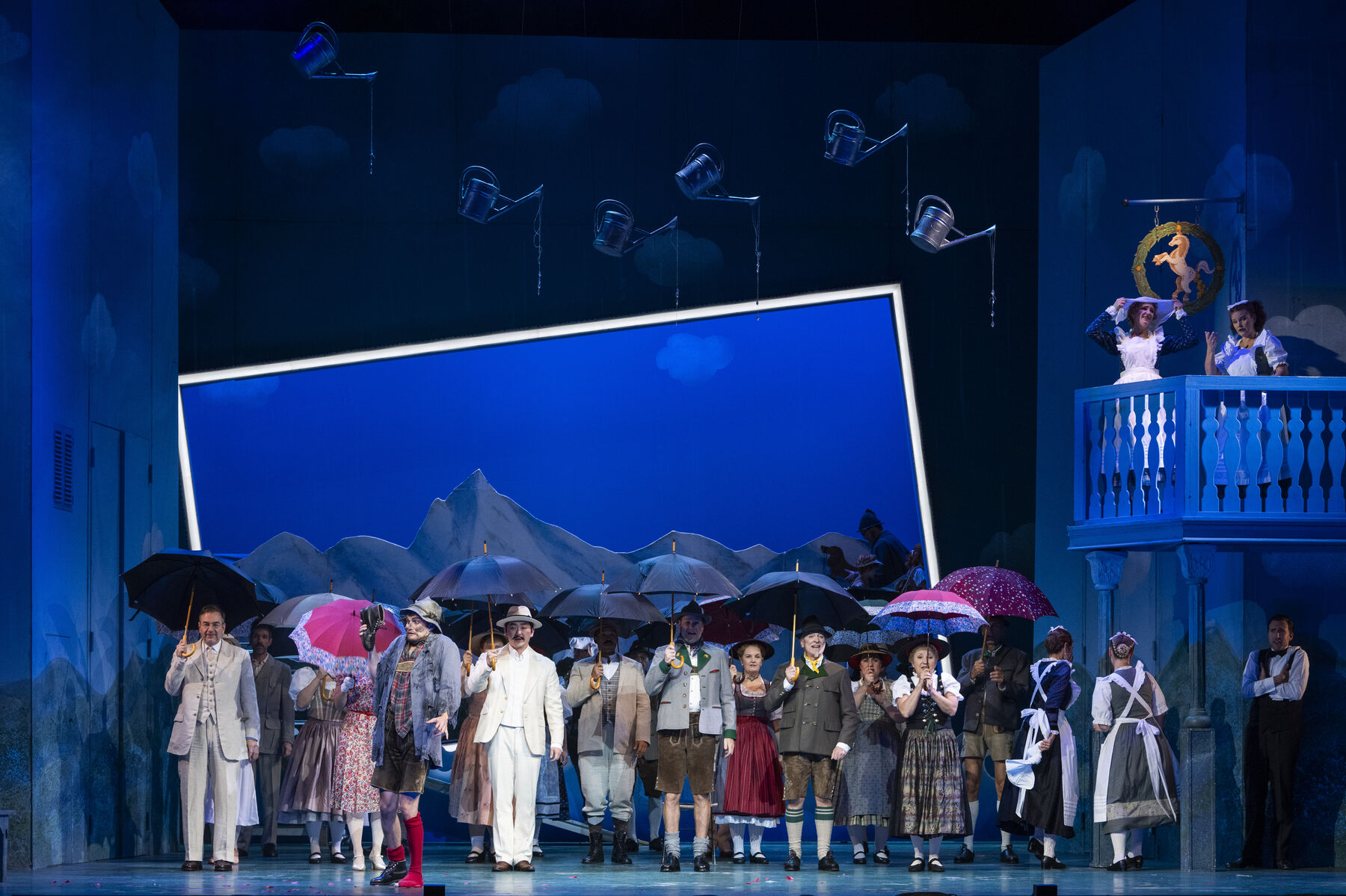 Das Ensemble steht mit Regenschirmen auf der Bühne. Gießkannen hängen an der Decke. Das Bühnenbild ist blau.