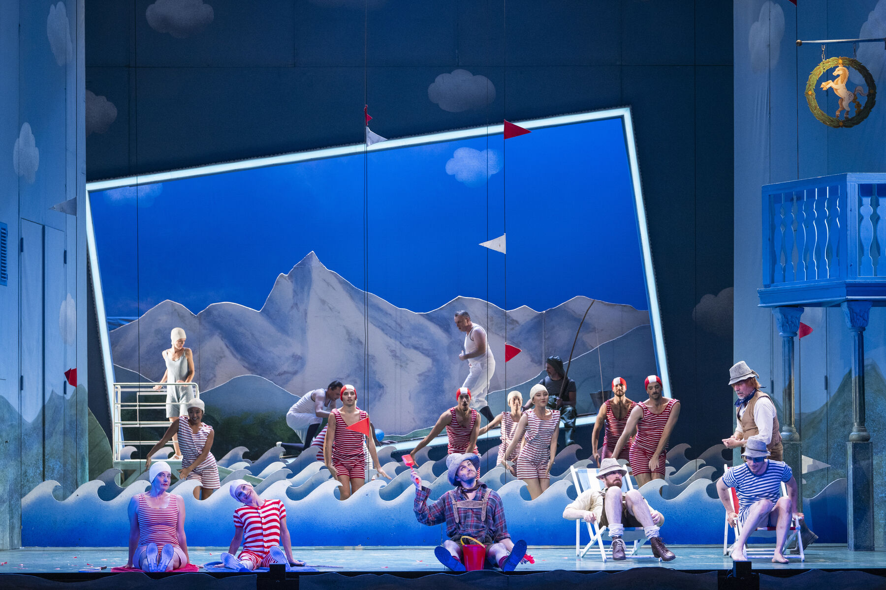 Ein Darsteller und da Ballettensemble stehen und sitzen auf der Bühne. Es sind Wellen zu sehen, sie tragen badekleidung.