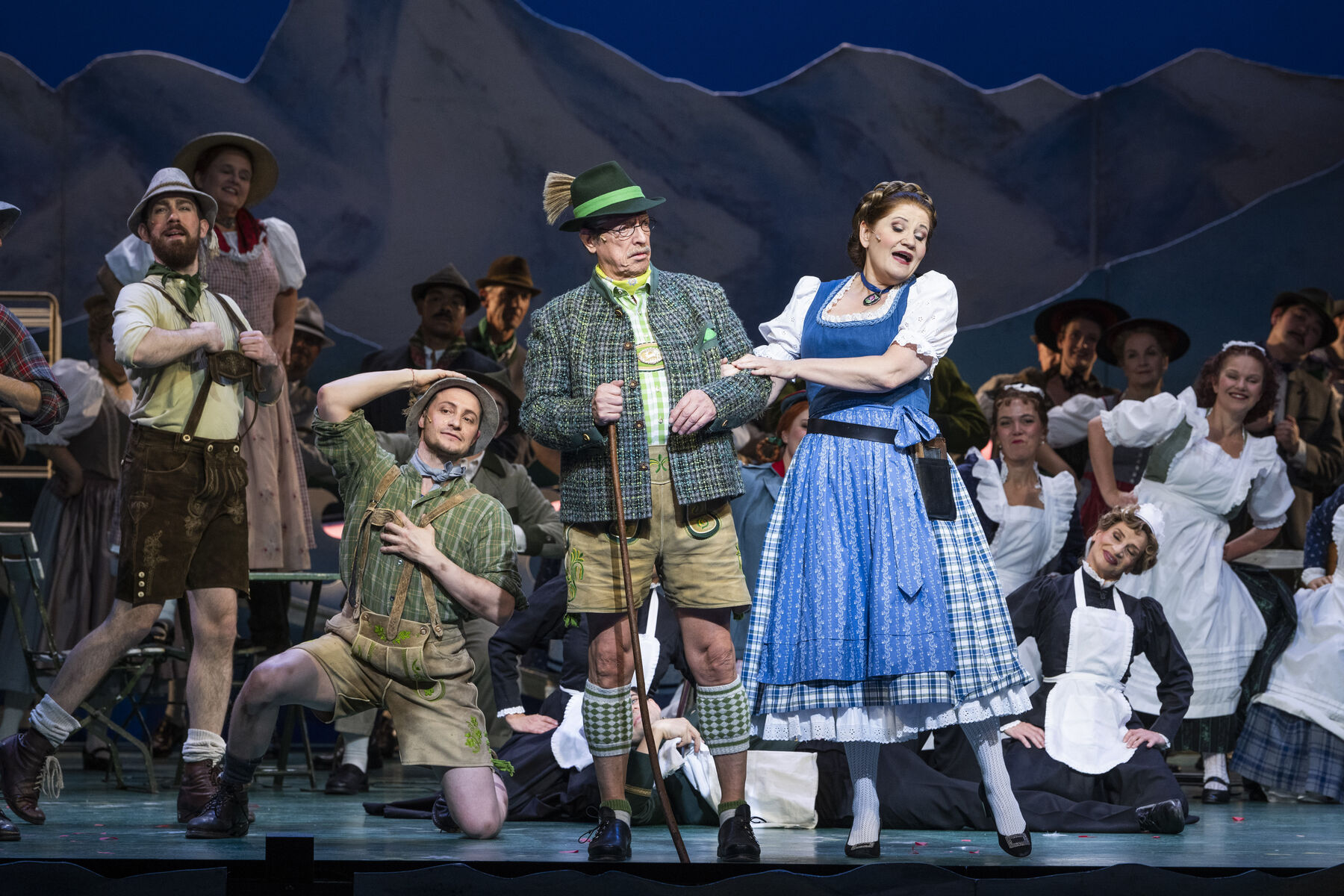 Drei Männer und eine Frau stehen in Tracht auf der Bühne. Hinter ihnen sind weitere Mitglieder des Ensembles zu sehen.