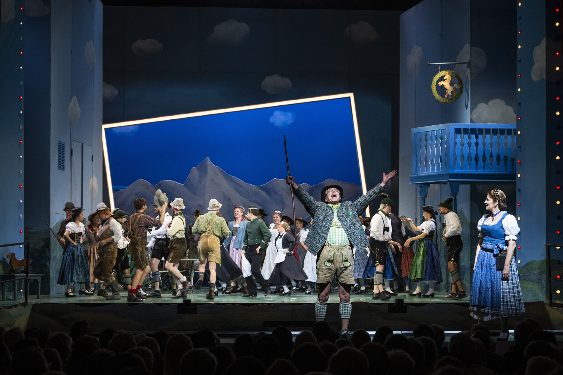 Auf der Bühne sieht man die Darstellerinnen. Davor kann man einen Mann und eine Frau in Tracht sehen. Er streckt seine Arme in die Luft.