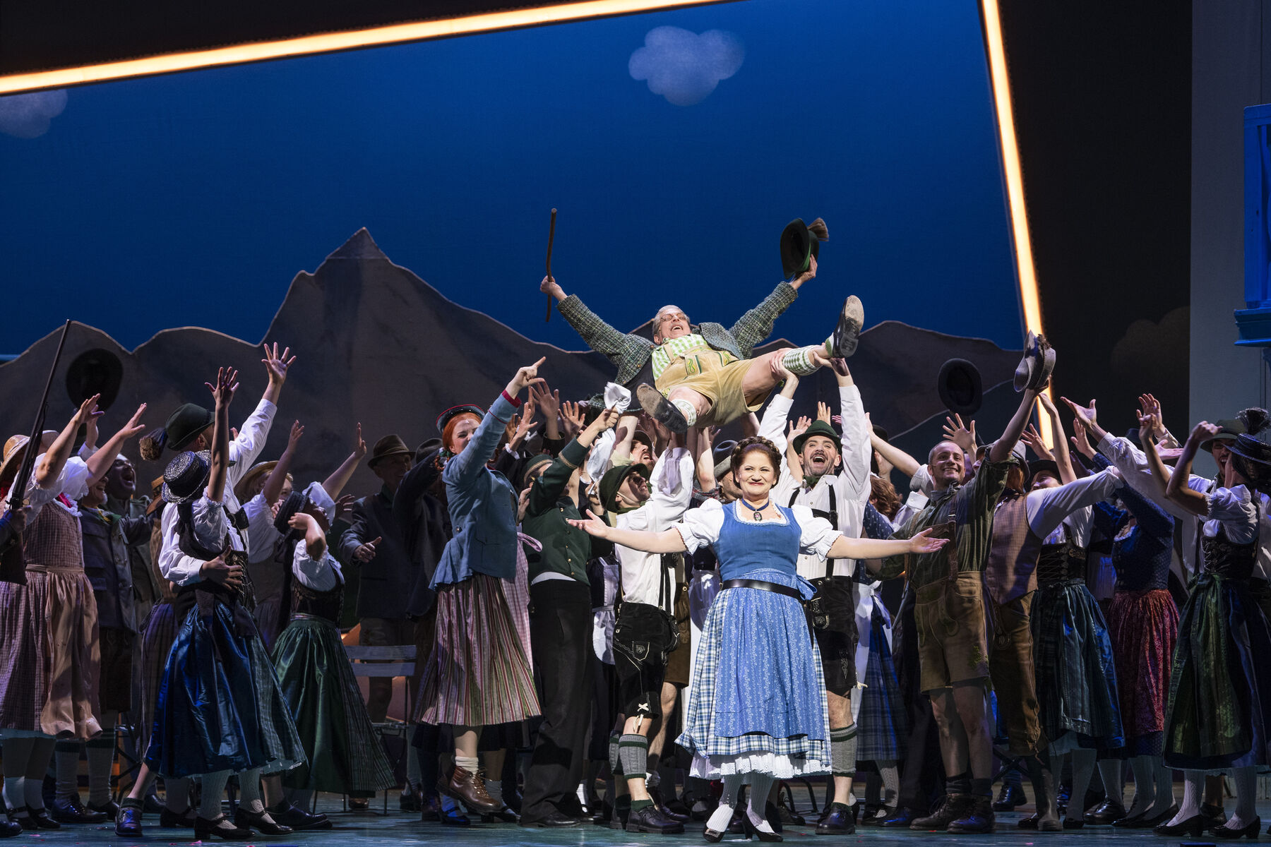 Das Ensemble ist auf der Bühne zu sehen, sie strecken die arme in die Luft. Manche tragen einen Mann. Eine Frau steht vor allen.