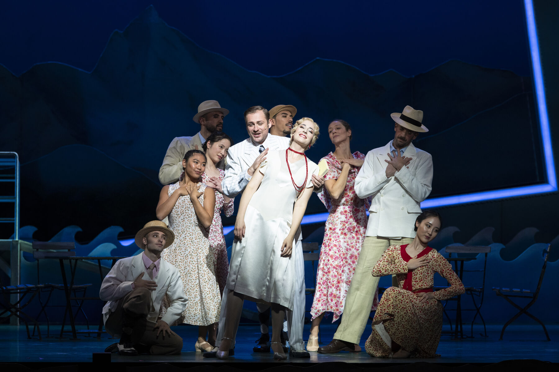 Zwei Darstellerinnen, weiß gekleidet, stehen auf der Bühne. Mitglieder des Balletts steht um sie herum.