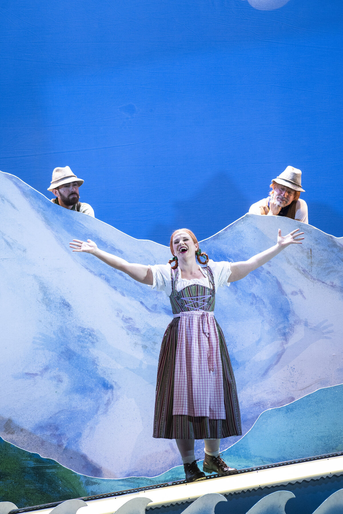 Eine Frau im Dirndl steht im ausgestreckten armen auf der Bühne und singt. Dahinter sind Berge zu sehen und zwei Männer welche sie anschauen.