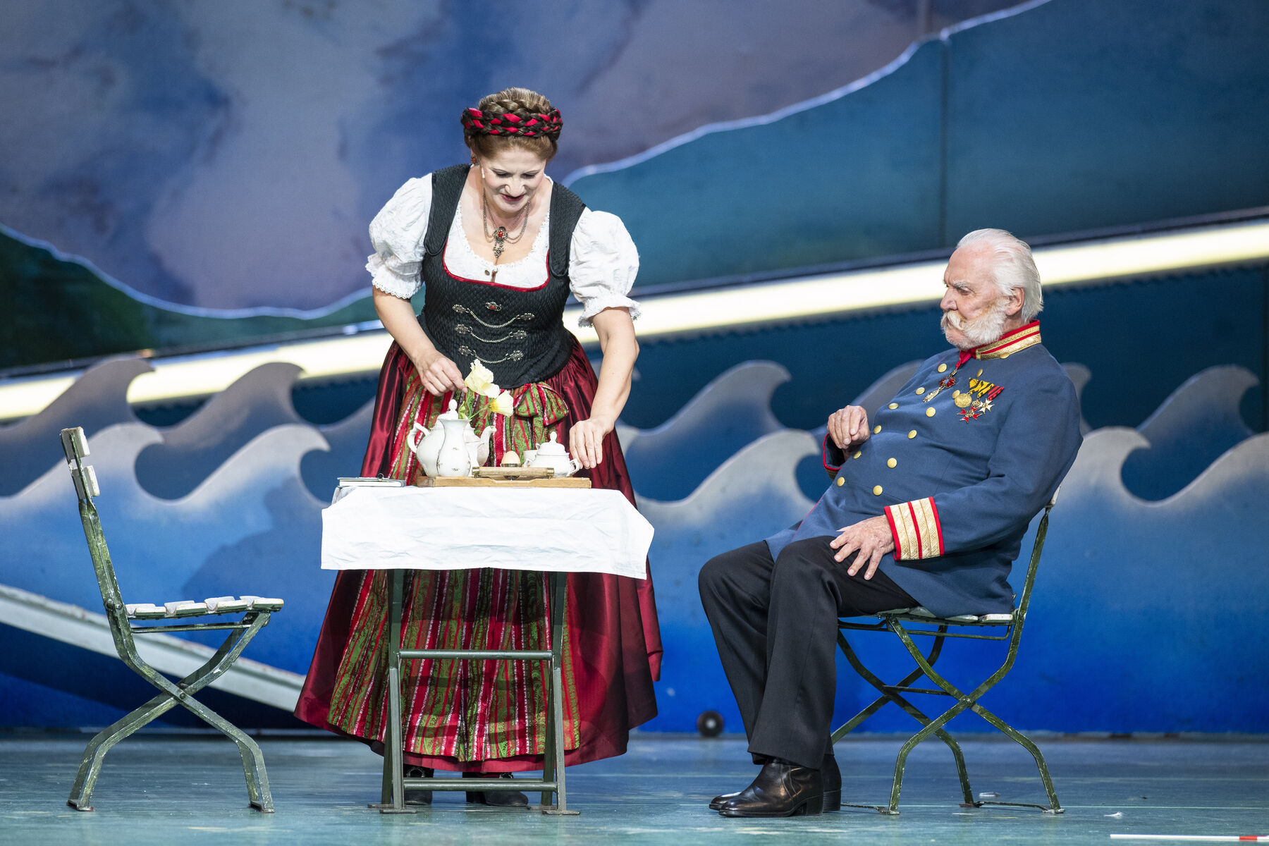 Eine Frau im Dirndl steht hinter einem Tisch. Auf einem der Stühle sitzt ein Mann als Kaiser gekleidet.