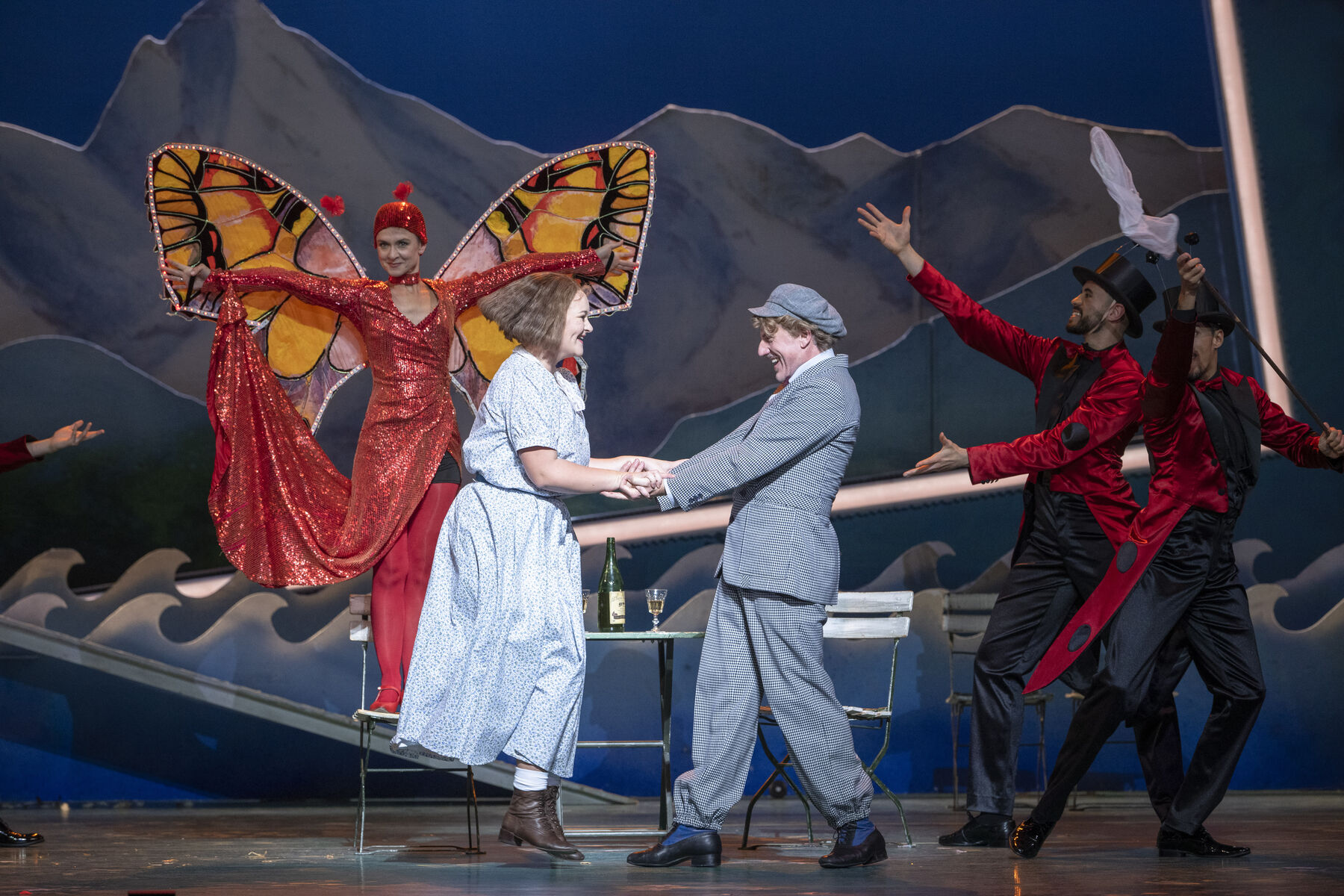 Eine frau und eine Mann halten sich an den Händen fest. Dahinter sieht man Mitglieder des Ballettensembles in rot.