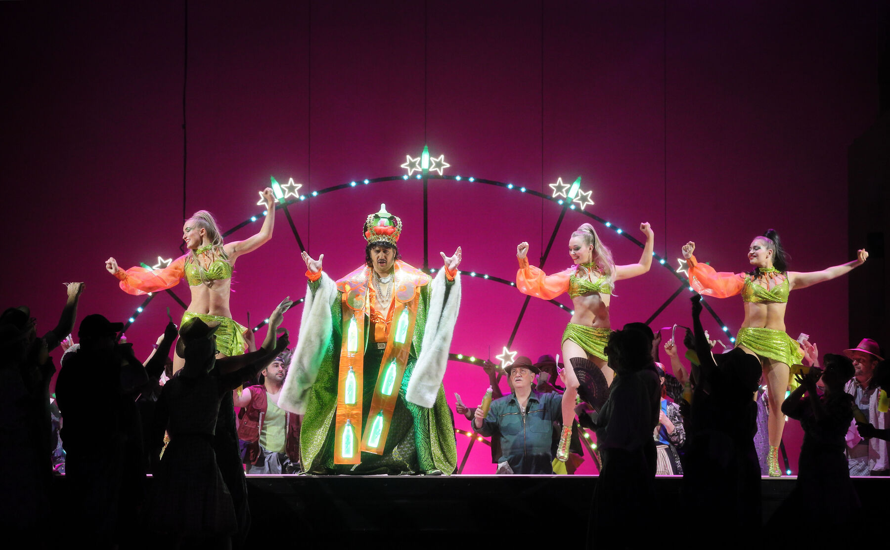 Dulcamara steht mit drei Showgirls auf einer Bühne. Um sie herum ein begeistertes, nicht beleuchtetes Publikum. Dulcamara trägt ein glitzerndes, orange-grünes kaiserliches Krönungsgewand. Im Hintergrund ist ein Metallgestell, das an ein halbes Spinnennetz erinnert, er ist violett beleuchtet.