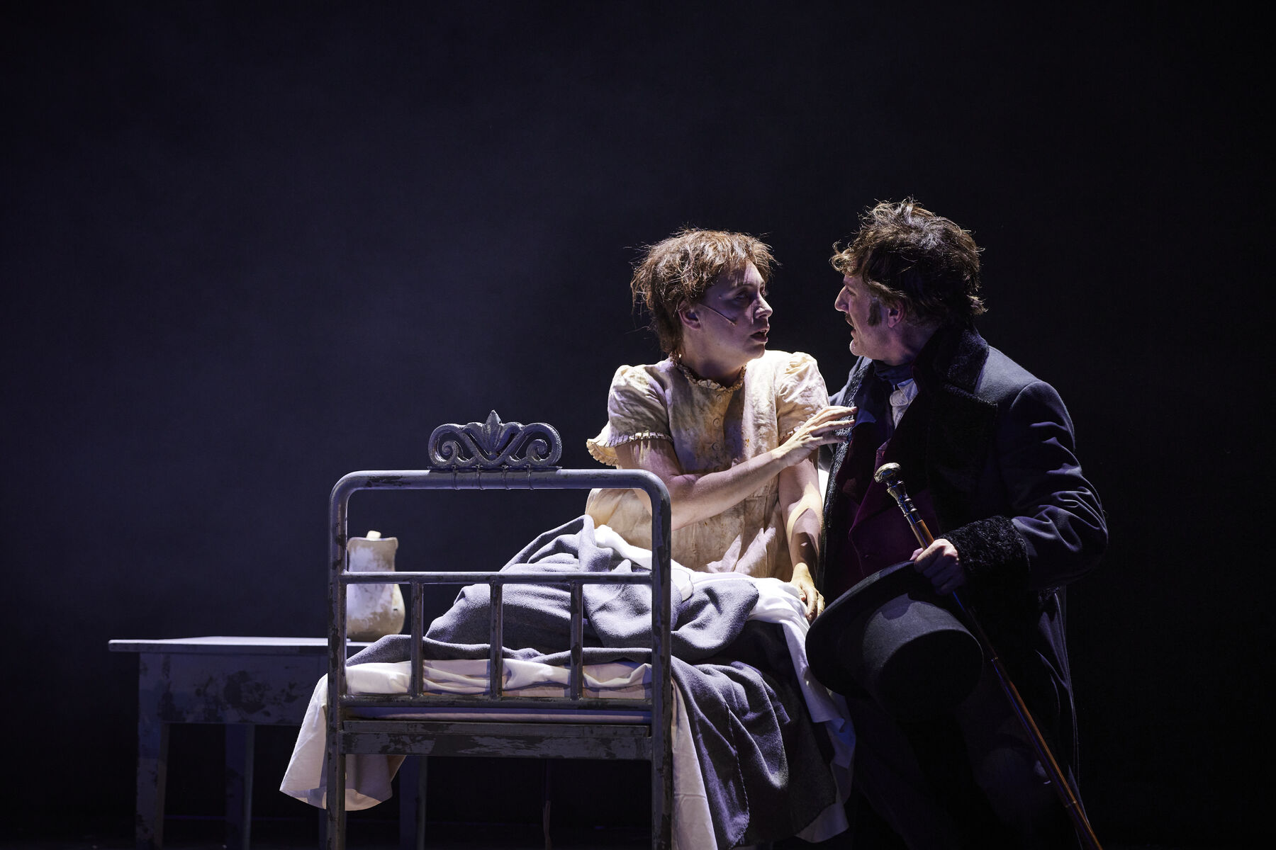 Auf einem alten Bett mit Nachttisch links daneben sitzen Fantine in hellem Nachtkleid und Jean Valjean rechts von ihr. Die beiden schauen sich an. Der Hintergrund ist schwarz.