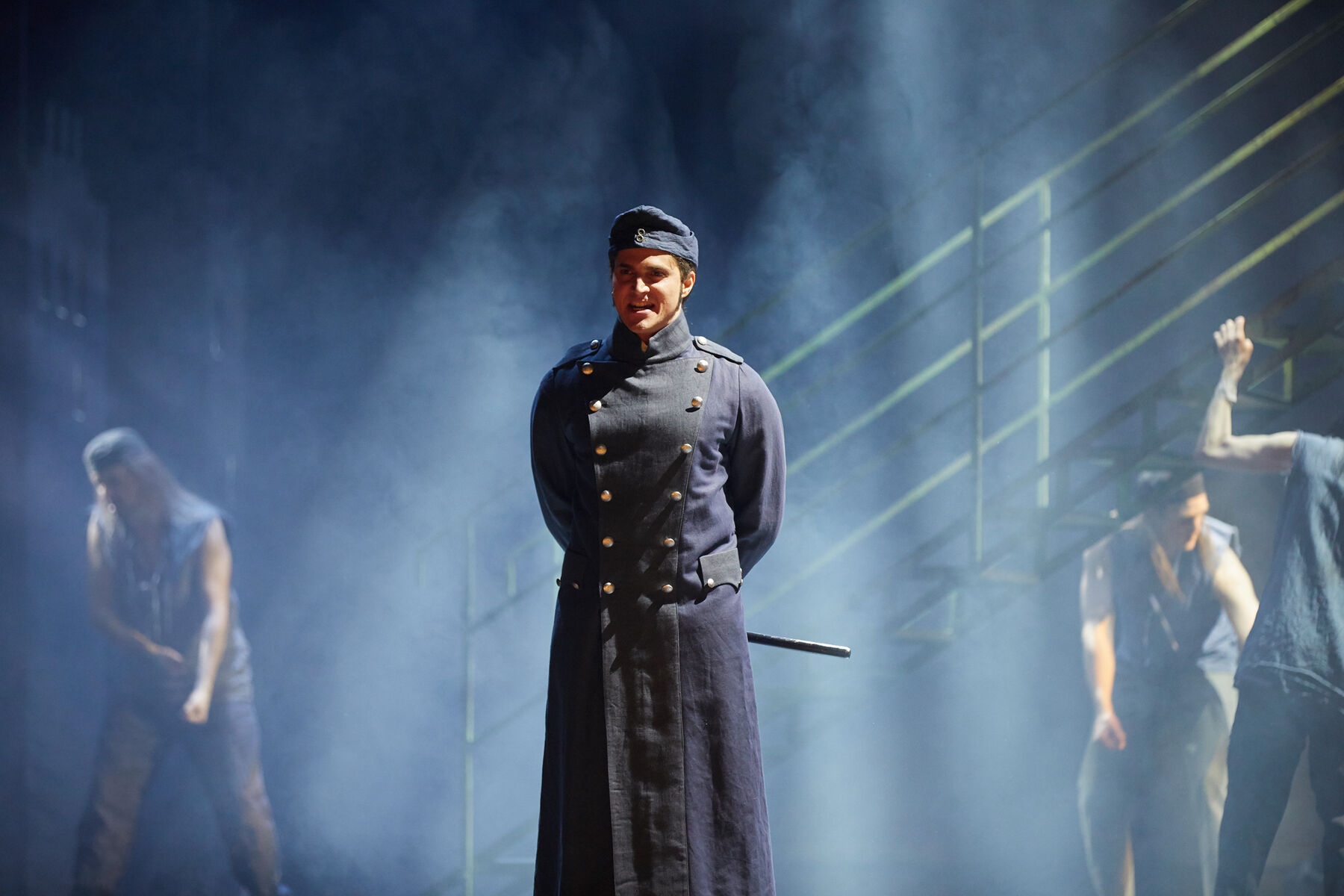 Javer steht in dunkel blauer Uniform, singend auf der Bühne. Im Hintergrund sieht man eine Treppenaufgang und Mitglieder des Ensembles. Es ist nebelig.