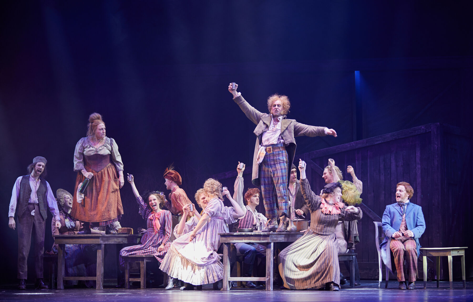 Auf Holztischen und -bänken sitzt das Ensemble und feiert ausgelassen. links auf einem der Tische steht Madame Thénardier mit einer Weinflasche in der Hand sie schaut zu Thénardier welcher rechts auf einem der Tische steht und feiert. Alle tragen Arbeiterkleidung.
