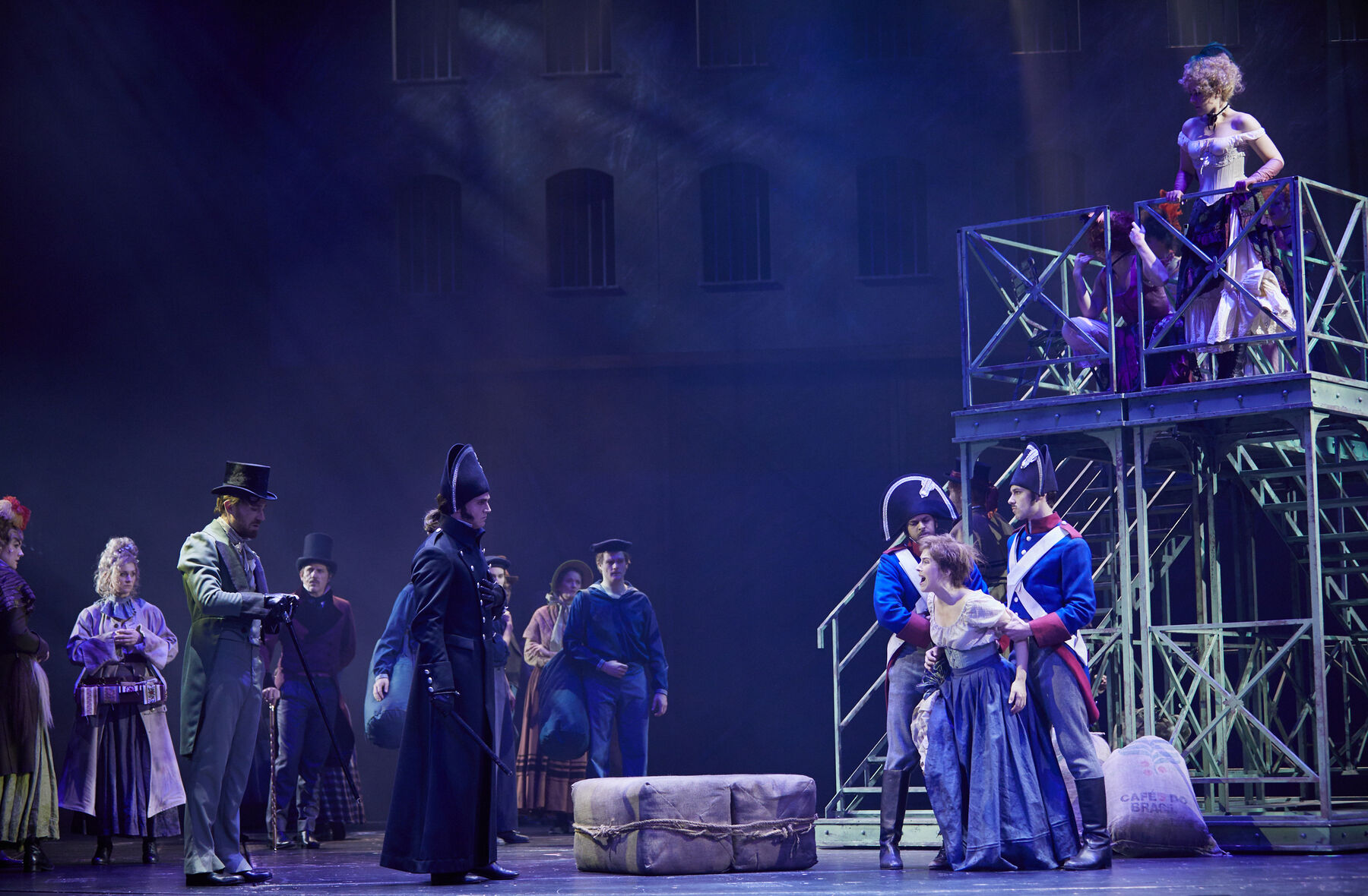 Auf der Bühne sieht man links Javert in uniform, ihm gegenüber mit etwas Abstand steht Fantine in einem Kleid, sie wird festgehalten. Das weitere Ensemble steht verteilt auf der Bühne. Auch auf der Treppe, am rechten Bildrand,  welche oben eine Plattform besitzt, stehen Ensemble Mitglieder. Im Hintergrund sieht man ein Gebäude.