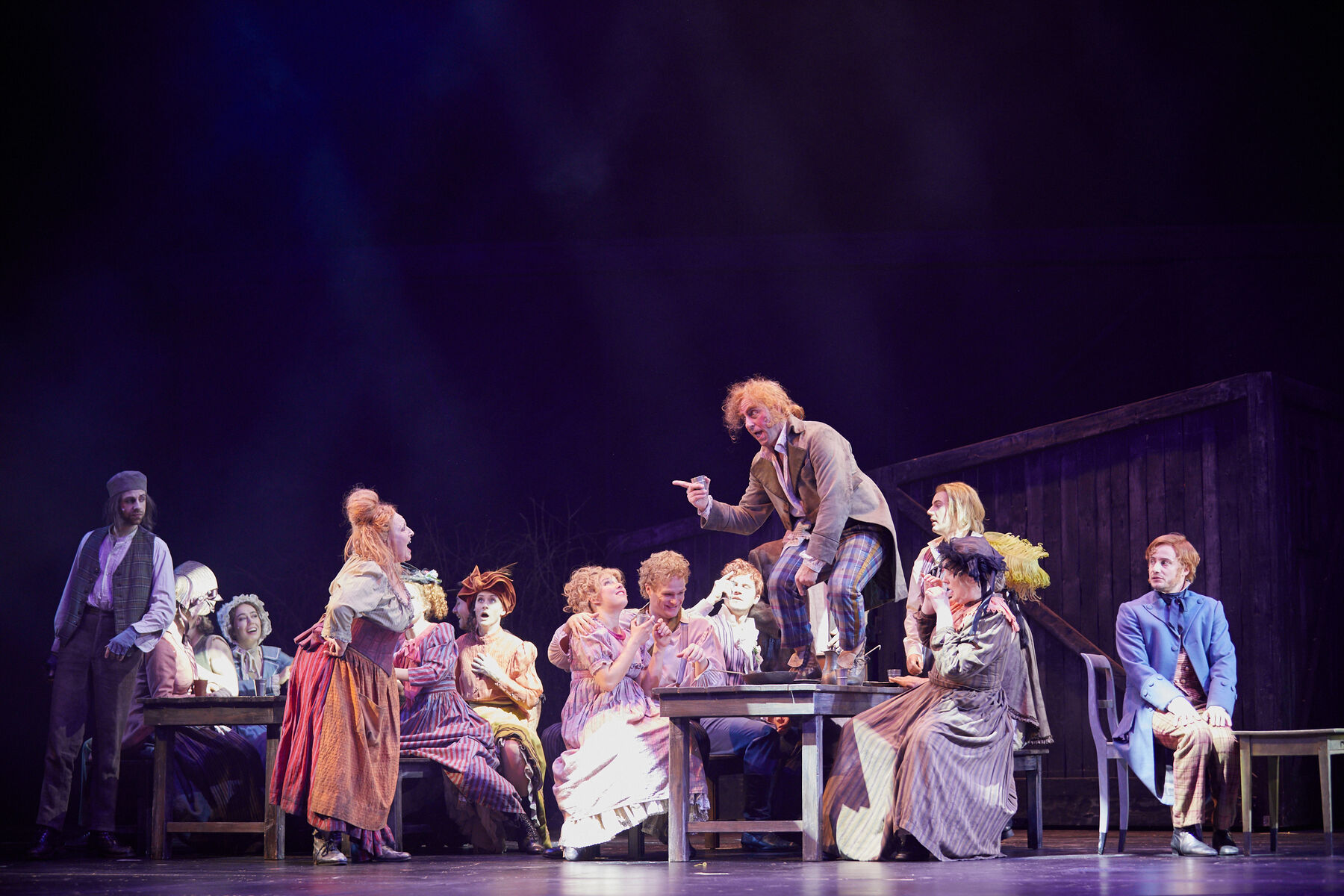 Auf Holztischen und -bänken sitzt das Ensemble und feiert. Links auf der Bühne steht Madame Thénardier, sie singt und schaut zu Thénardier welcher rechts auf einem der Tische steht und sich zu ihr bückt und mit dem Finger auf sie zeigt. Alle tragen Arbeiterkleidung.