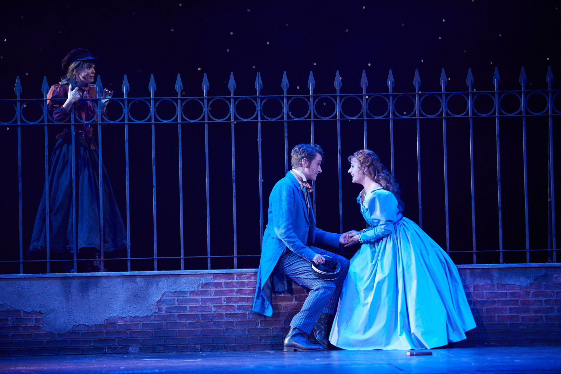 Cosette und Marius sitzen auf einer mauer, sie halten ihre Hände und schauen sich an. Dahinter ist ein Zaun. Eine junge Frau schaut darüber.
